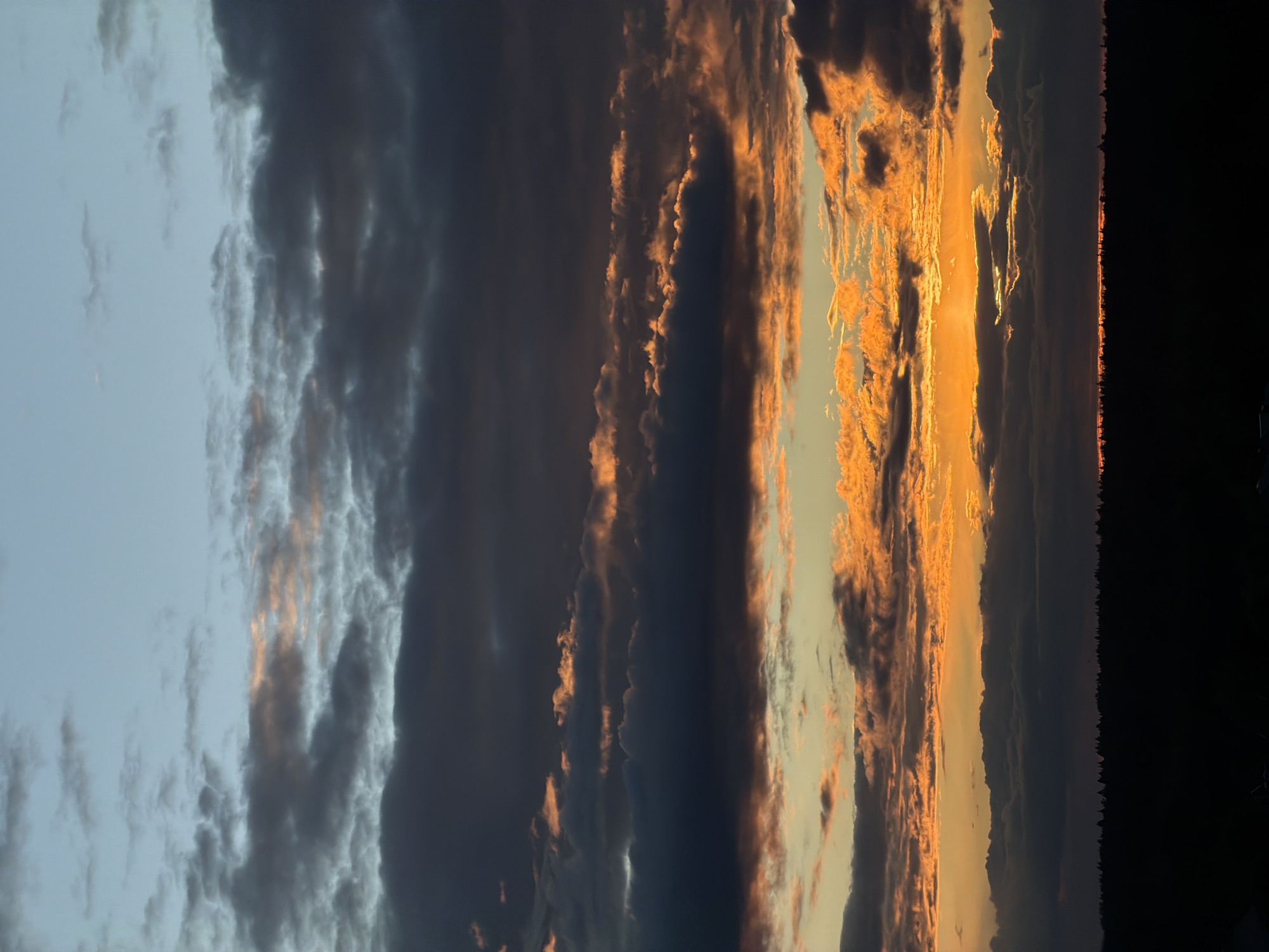 A photograph captures a landscape during twilight. Dark and ominous clouds are spread out. The horizon is a dark silhouette, possibly trees. There is no evidence of people. The location cannot be determined from the image.