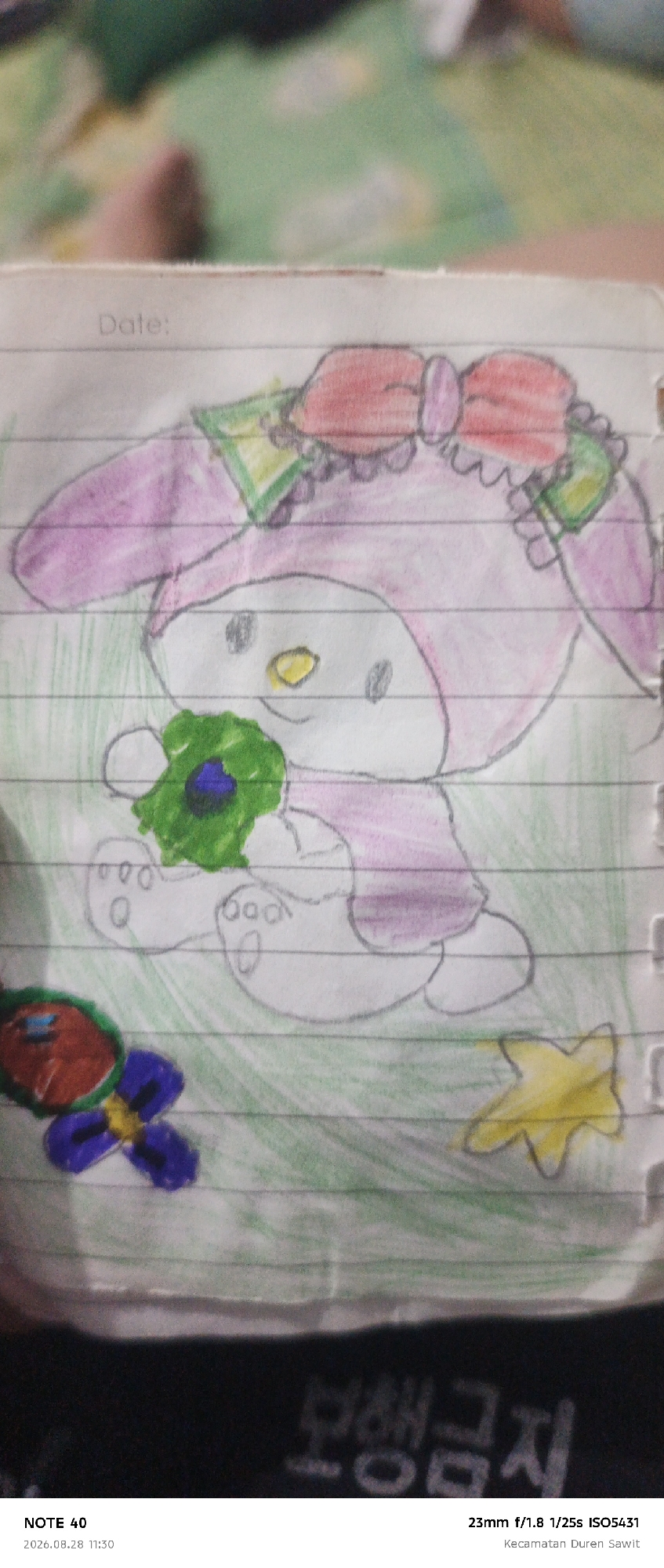 The image is a close-up, slightly blurry, overhead shot of a hand-drawn picture on lined paper. The drawing depicts a cartoon character, likely My Melody, colored in pink and purple, with a red bow on its head. The character is holding a green object with a dark blue center, possibly a flower or a ball. To the left of the character is a drawing of a red and blue object with a green center, and to the right, a yellow star. The background of the drawing is lightly sketched in green. The paper is placed on a dark surface. At the bottom of the image, there is text that reads "NOTE 40 2026.08.28 11:30" and "23mm f/1.8 1/25s ISO5431 Kecamatan Duren Sawit". The text "Kecamatan Duren Sawit" indicates the location.