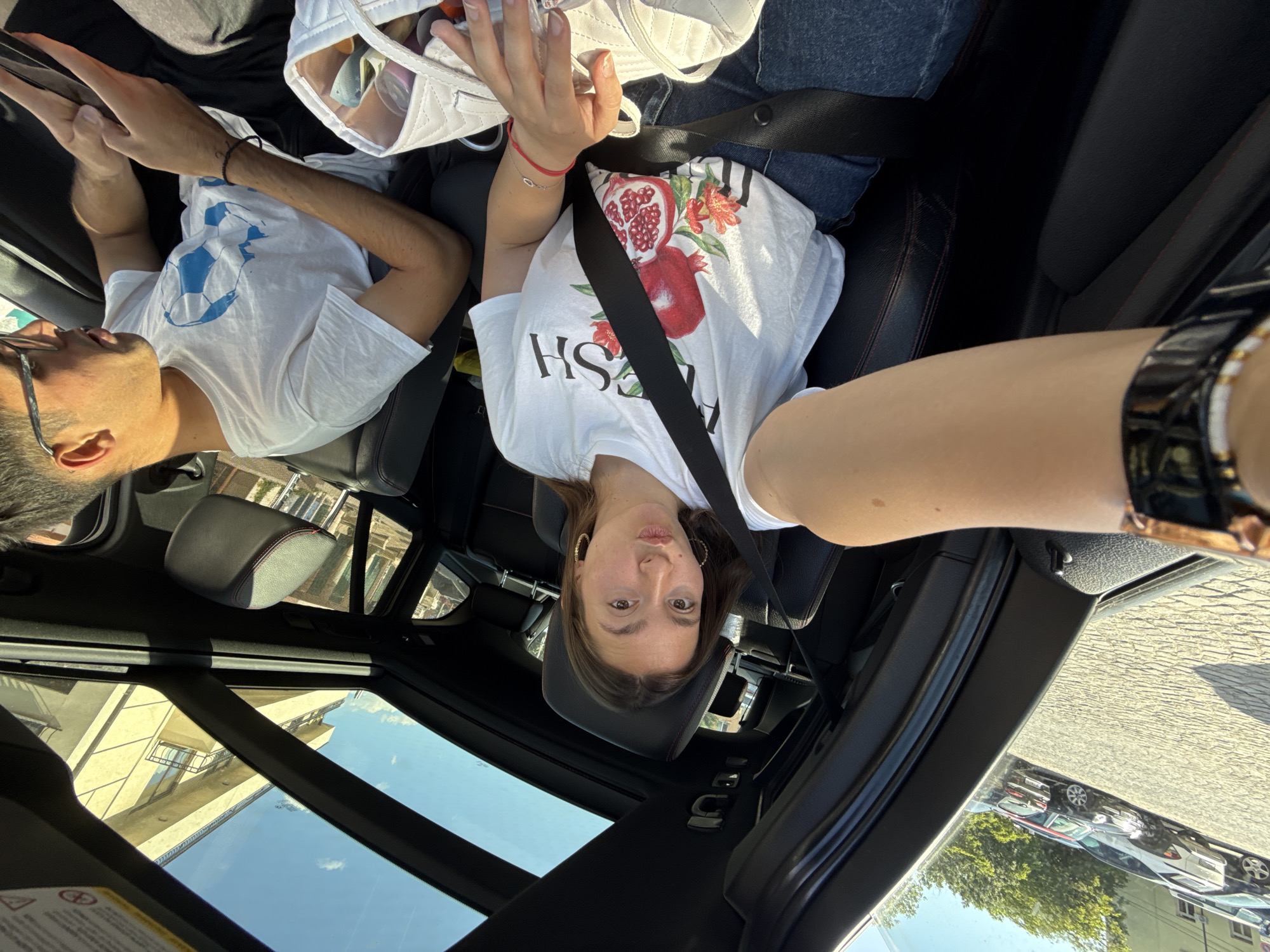 In Sofia, Bulgaria, on a sunny day, two young adults are in the front seats of a car. The woman, in the driver's seat, takes a selfie with her phone, puckering her lips. She wears a white t-shirt with a pomegranate design and blue jeans. A black seatbelt is across her chest. Beside her, the man in the passenger seat, wearing a white t-shirt with a blue soccer ball design and black shorts, looks down at his phone. A white quilted bag rests on their laps. The car's sunroof is open, revealing a clear blue sky and a building with balconies. Outside, cars are parked on a cobblestone street.