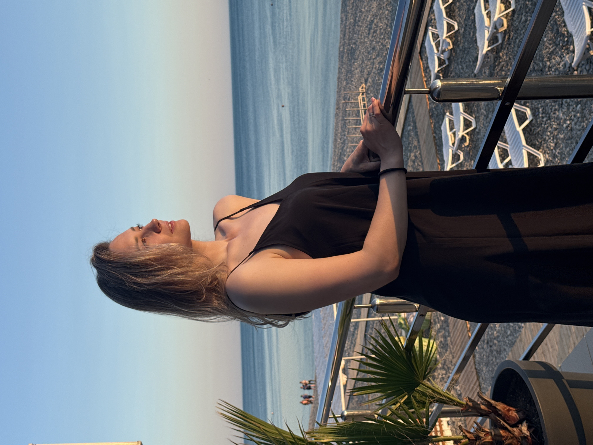 An outdoor scene in Nebug, Russia, depicts a young adult female standing on a balcony overlooking a pebble beach and the sea during the golden hour. The woman, with shoulder-length blonde hair illuminated by warm light, is positioned in profile, facing towards the right side of the frame. She wears a dark, possibly black, spaghetti-strap dress and rests both hands on a polished metal railing that spans the width of the balcony. A silver ring is visible on the ring finger of her left hand. To the immediate left of the woman, a large potted plant with green, palm-like fronds is visible. Beyond the railing and balcony floor, which appears to be dark gravel or pavement, a vast expanse of light grey pebble beach extends towards the tranquil blue-grey sea. Numerous unoccupied white plastic sun loungers are arranged in orderly rows across the beach. In the distance, near the waterline on the left, a small group of approximately four to five indistinct figures can be observed walking. The sky above the sea presents a gradient from a clear, light blue at the zenith to a lighter, more yellowish hue near the horizon, reflecting the warm, low-angle sunlight. Small red buoys are discernible on the surface of the calm sea.