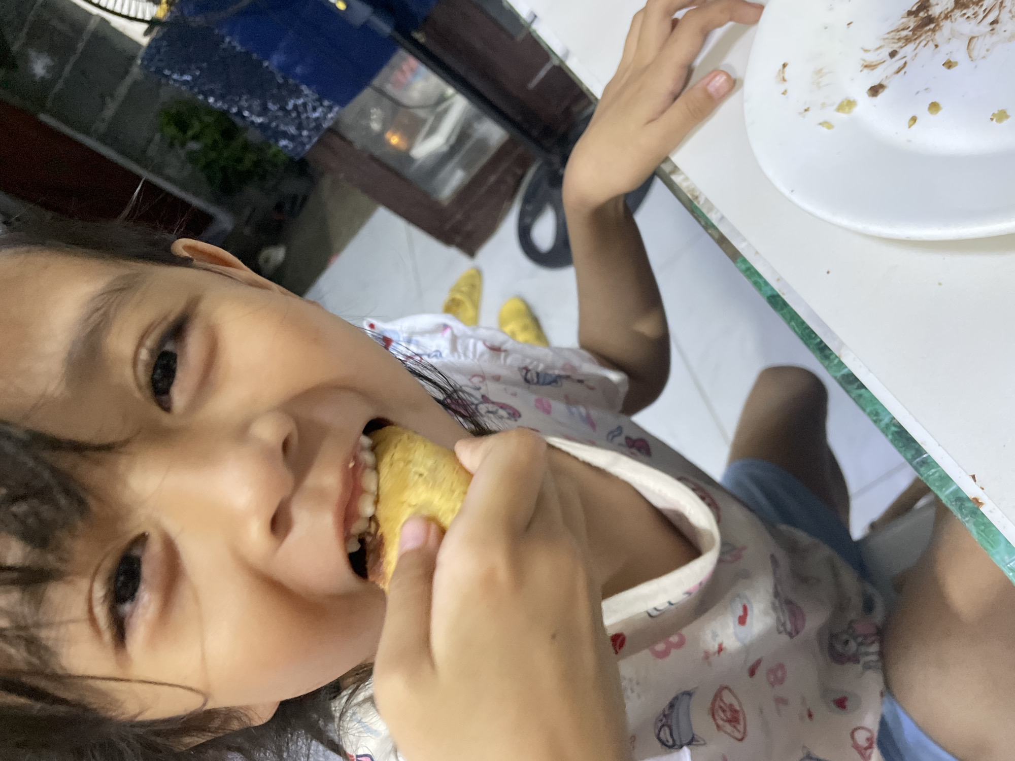A young girl is eating a piece of food, possibly cake, in what appears to be a residential dining area. She is wearing a patterned shirt. A white plate with crumbs is visible on the table. The location is indicated as Davao City, Philippines. There are yellow shoes on the floor.