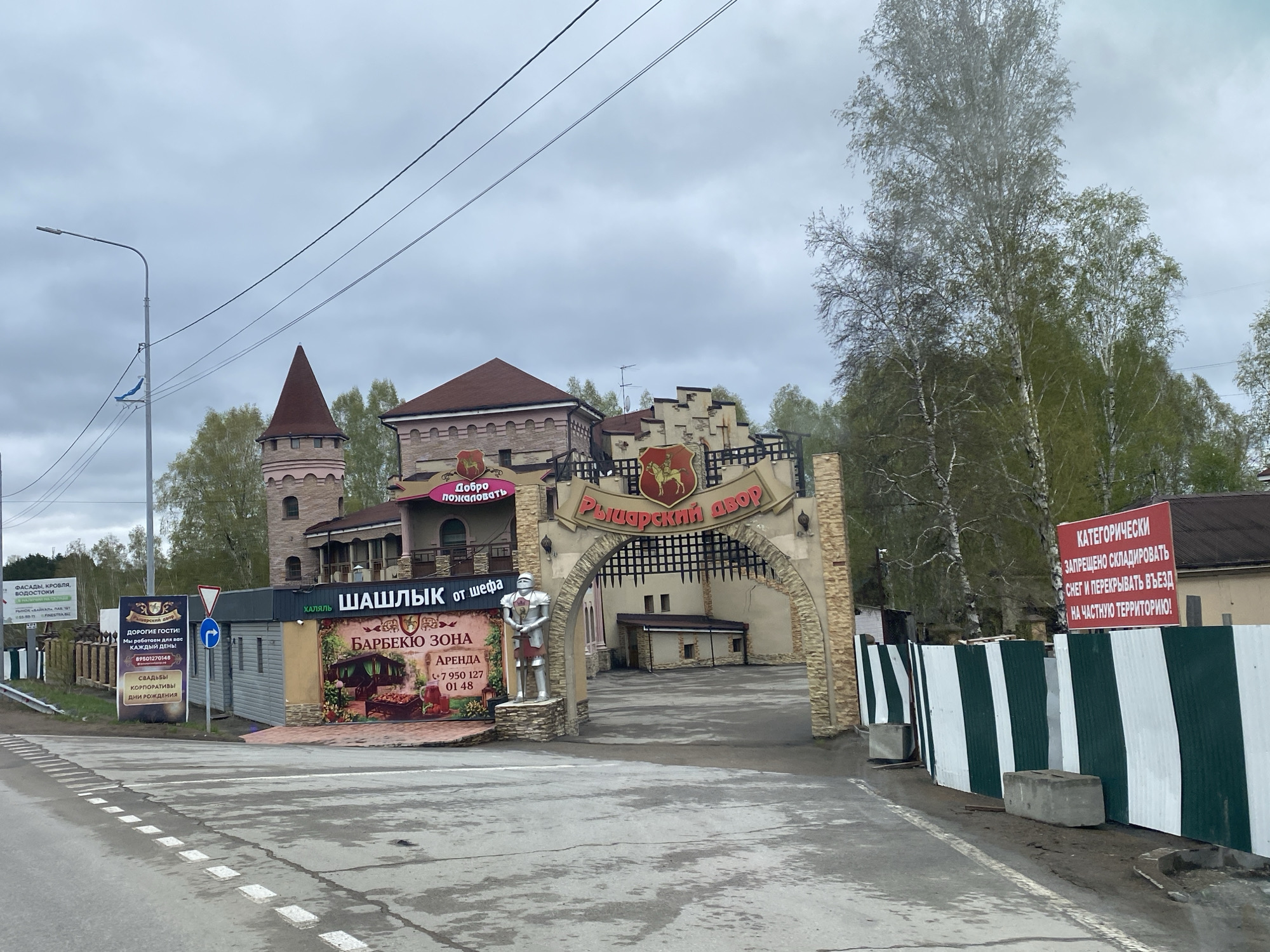 The image depicts a street scene in the city of Молодёжный, Russia, showcasing the entrance to a complex called "Рыцарский двор," which translates to "Knight's Courtyard." The entrance features a stone archway with an inscription in Russian. To the left of the entrance, there is a sign promoting a "BBQ zone" ("БАРБЕКЮ ЗОНА") offering rental services, along with contact information. There's also a smaller building with the words "ХАЛЯЛЬ ШАШЛЫК от шефа," indicating halal grilled meat from the chef.
A statue of a knight stands near the entrance. To the left of the statue, there are signs advertising construction materials (roofs, facades, etc.) and events (weddings, corporate events, birthdays). On the right side, a sign warns against blocking the entrance with snow, stating that it is private territory. A fence of green and white vertical stripes is visible next to the sign.
In the background, a building resembling a castle is visible, with a tower and brickwork. The sky is overcast, suggesting a cloudy day. The road in the foreground is wet, indicating recent or ongoing precipitation. A utility pole with wires is on the left side of the street.