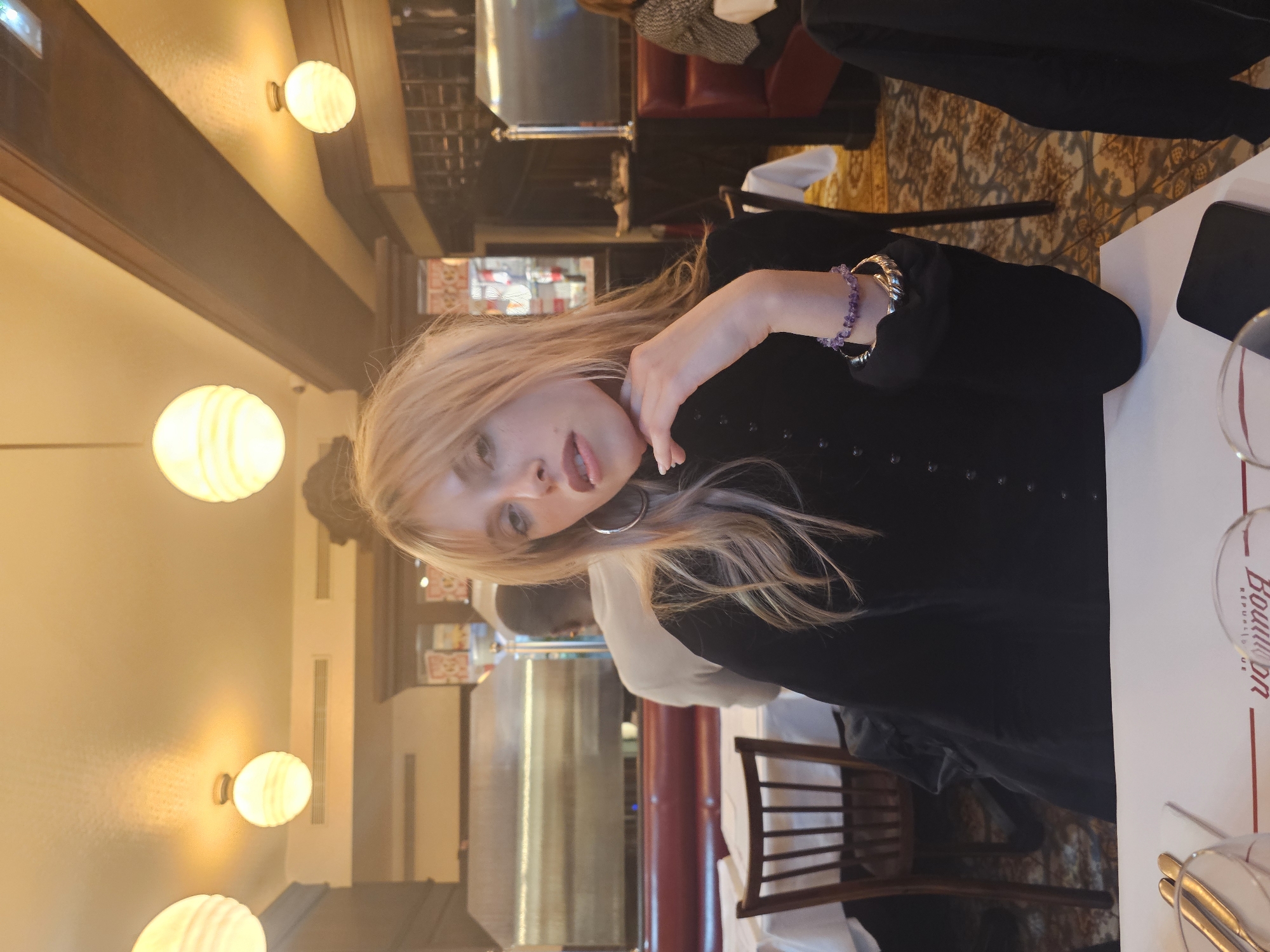 A medium-shot, rotated image features a woman with blonde hair and fair skin, wearing a black, button-down shirt. She is positioned at a table inside what appears to be a restaurant or brasserie. A bracelet adorns her left wrist. The table is set with glassware and silverware, and a mobile phone is placed on it. The logo of "Bouillon République," a well-known Parisian restaurant, is visible on the table. In the background, other patrons and restaurant furnishings, like chairs and tables, are visible. The overall lighting suggests a warm, indoor environment. The scene appears to be taking place in Paris, France, specifically at Bouillon République, based on the logo and general ambiance.