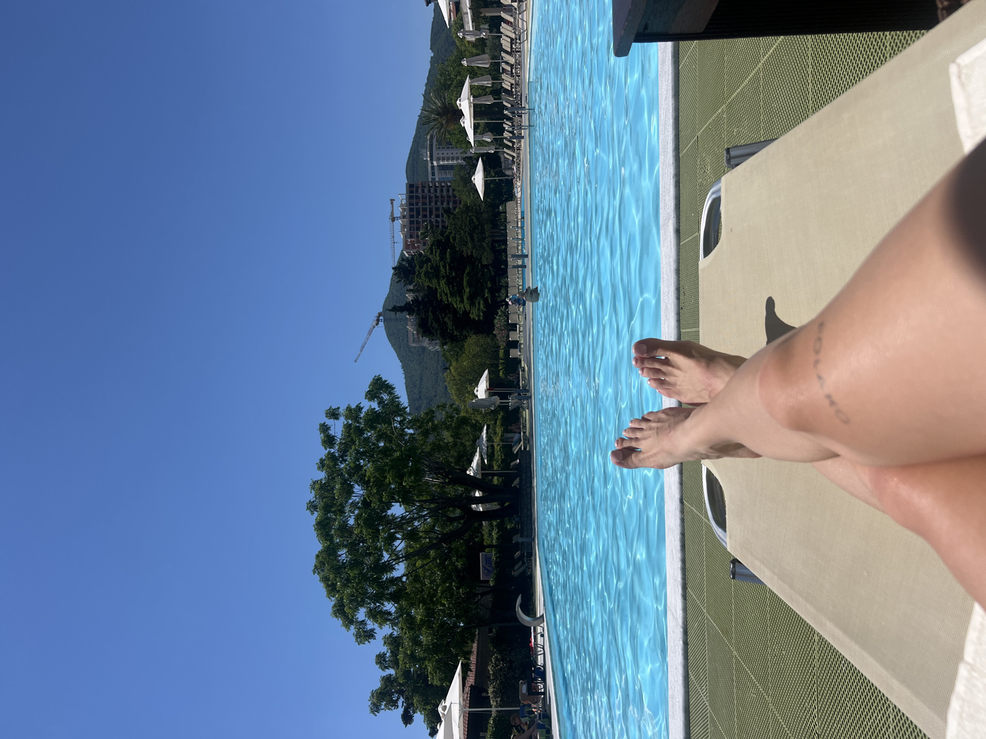 A daytime, outdoor shot depicts a person reclining on a beige lounge chair, positioned next to a swimming pool with light blue water. The person's legs and feet are visible, suggesting they are relaxing by the pool. The pool is lined with a white edge and bordered by a green textured surface. The background shows a landscape with trees, a building under construction, a mountain, and a clear blue sky. White parasols and lounge chairs are visible along the pool's edge. Based on the landscape features, the location is likely in or near Budva, Montenegro.