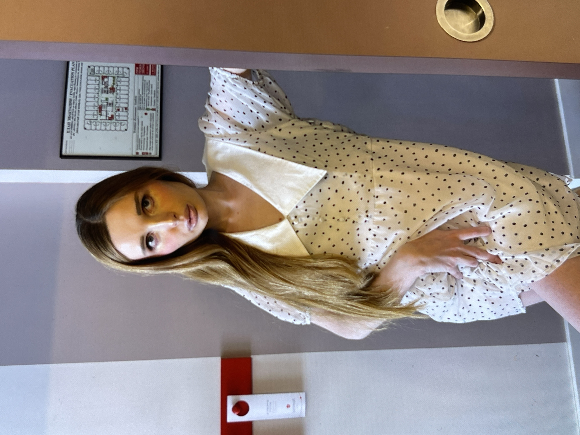 A young woman with long, light brown hair and a white dress with black polka dots stands leaning against a doorframe. Her face is partially obscured, but makeup is visible, with prominent blush and eyeshadow. In the background, an evacuation plan is displayed on a wall, and a white dispenser with a red top is mounted on a red bar. The environment appears to be indoors, possibly a hotel room hallway, with a light gray wall on the left and a brownish doorframe on the right.