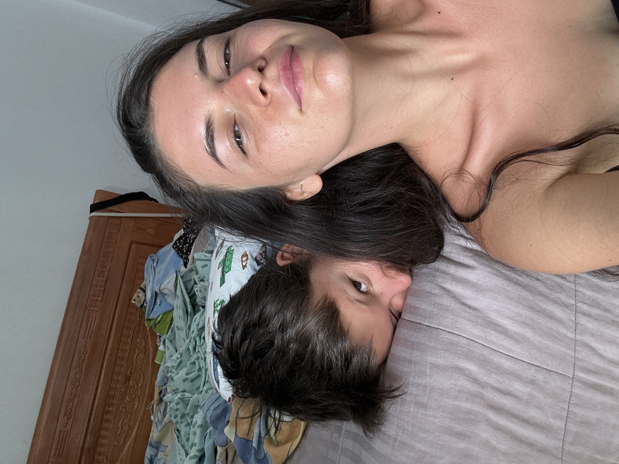A portrait of a young woman and a young man lying on a bed with light gray bedding. The woman has long dark hair, fair skin, and is looking directly at the camera. The man has short dark hair and is lying slightly below her, also looking at the camera. They are positioned head-to-head. In the background, there is a wooden door and a pile of colorful, patterned fabric or clothes. The location is in a room and possibly a hotel in the city of Mũi Né, Vietnam.