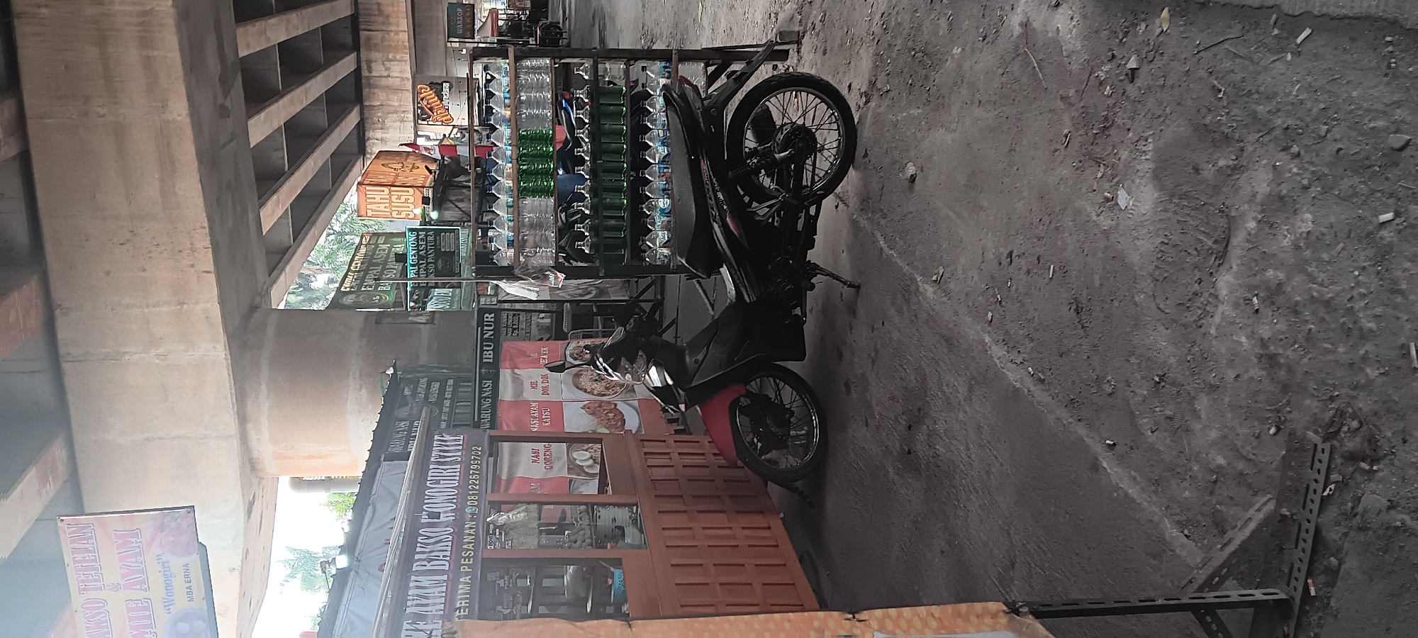 A motorcycle is parked in front of a food stall under an overpass in South Jakarta, Indonesia. The stall has a sign that reads "AYAM BAKSO MONOGIRI STYLE" and offers dishes like "NASI GORENG" and "MIE AYAM". A rack of bottled drinks, including water and what appears to be cooking oil or syrup, is located next to the stall. The ground is paved with concrete and shows signs of wear and tear, with cigarette butts scattered around. The overpass structure is visible above, casting shadows on the scene. There are no people clearly visible in the image.