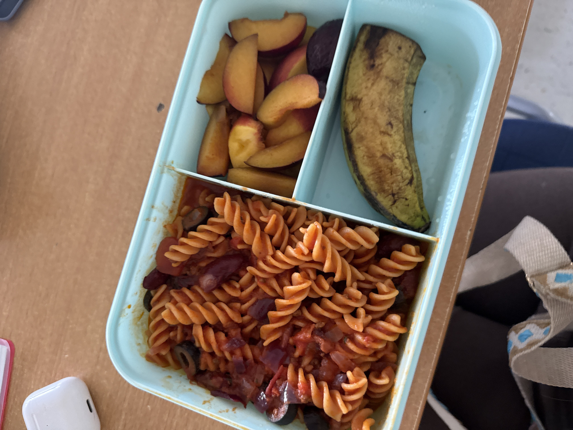 A light blue lunchbox sits on a wooden table. The lunchbox is divided into three compartments. The largest compartment on the left contains rotini pasta with a red sauce, olives, and what appear to be kidney beans and chopped red onion. The middle compartment to the right contains sliced peaches. The smallest compartment on the bottom right contains a whole, unpeeled plantain with some brown spots. In the background, to the left of the lunchbox, is a white wireless earbud case. The image does not contain any information that allows for a determination of the location in the city of Hanau, Germany.