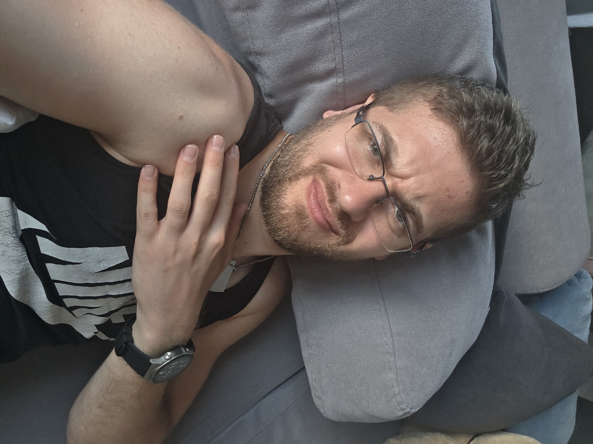 A male individual is depicted in a close-up, high-angle selfie, reclining on a grey fabric sofa or bed. He possesses short brown hair, a light brown beard, and wears dark-framed rectangular glasses. His facial expression conveys discomfort or mild displeasure, characterized by furrowed brows and slightly pursed lips. He is wearing a black tank top with a partially visible white "NIKE" logo on the chest. A silver-colored chain necklace with a rectangular pendant hangs around his neck. His left hand is positioned on his right shoulder/upper arm. A dark-colored smartwatch with a round face and black band is visible on his left wrist, displaying the time as "19:15". The background consists of multiple fabric pillows in shades of light grey, dark grey, and light blue. The scene is indoors, located in the city of Yagodovo, Bulgaria.