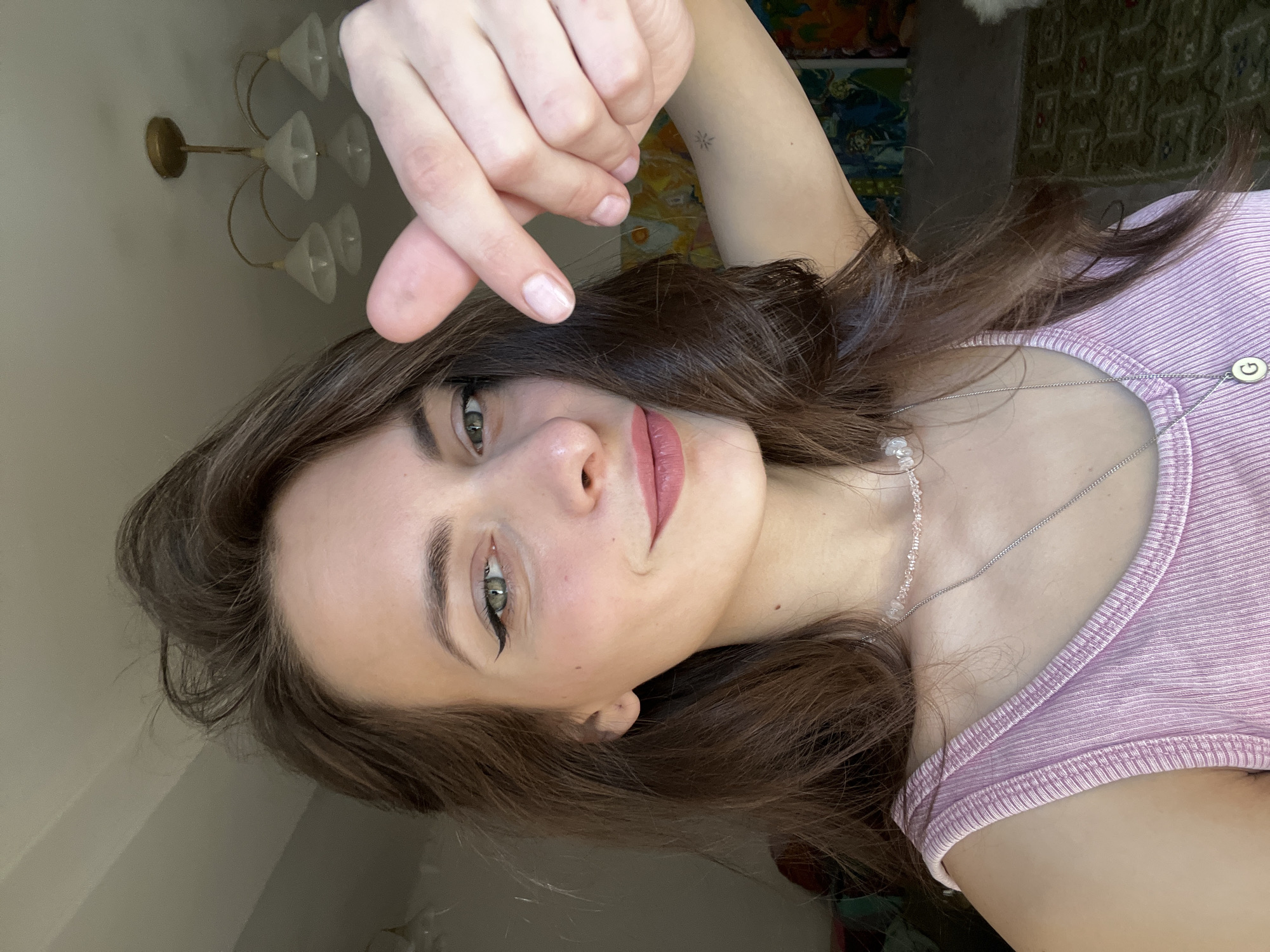 A woman with brown hair and light skin is in a selfie, holding her hand up, fingers crossed. She is wearing a pink ribbed top and a silver necklace with light-colored beads and a circular charm. Her eyes are green, and she is wearing black eyeliner. In the background, there is a chandelier and artwork. It is not possible to determine the exact location in Krasnoyarsk, Russia from this image.