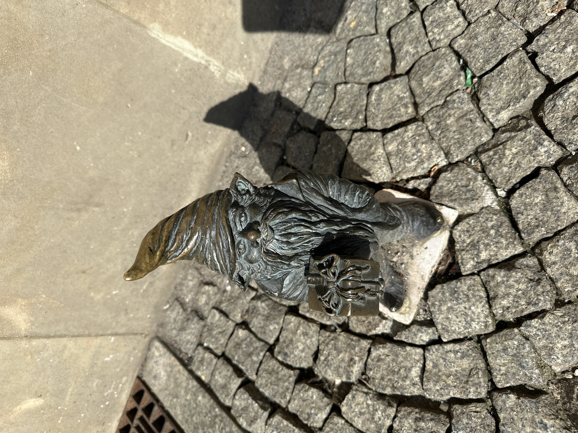 A bronze statue of a dwarf, known as a Wrocław dwarf, sits on cobblestone pavement in the city of Wrocław, Poland. The dwarf is wearing a long, pointed hat and has a long beard. He is holding a small, rectangular box with a bow on top, likely a gift. The cobblestone pavement surrounds the statue, and a concrete sidewalk is visible to the left. The statue casts a shadow on the concrete sidewalk. The lighting suggests a sunny day.