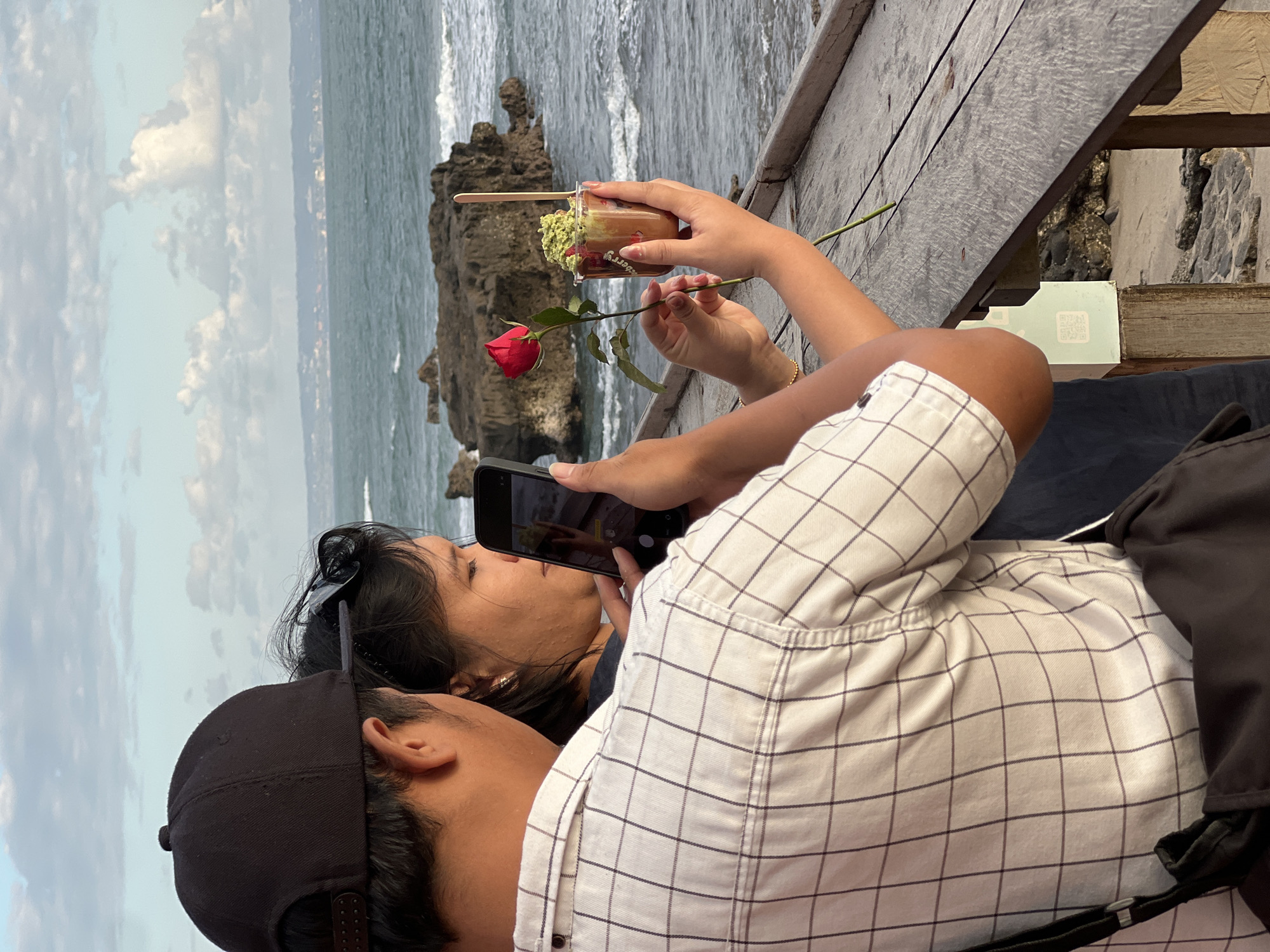 Two people are near the ocean. A person in a black baseball cap and a checkered shirt is taking a photo of a second person who is holding a red rose and a cup of food. The second person has long dark hair and sunglasses on their head. The ocean has waves, and there are rocks in the water. The location is likely a restaurant or cafe with a view of the ocean in North Kuta, Indonesia.
