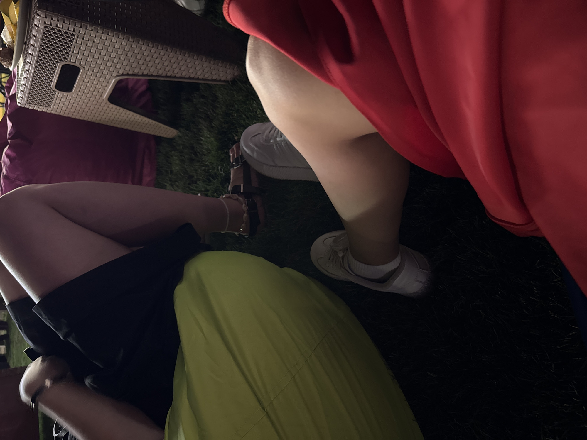 Two individuals are seated on the ground in an outdoor setting, likely a park or backyard, with grass visible beneath them. The person on the left wears a black skirt and is seated on a large, bright green beanbag. They have a bracelet on their wrist. The person on the right wears a red garment, possibly a dress or skirt, and is seated near a small, woven brown table. White sneakers are visible on both individuals. In the background, a large, pink beanbag is partially visible, as well as hints of other people and some food items on the table. The lighting suggests it may be evening or nighttime. The location is identified as Unknown, Egypt.