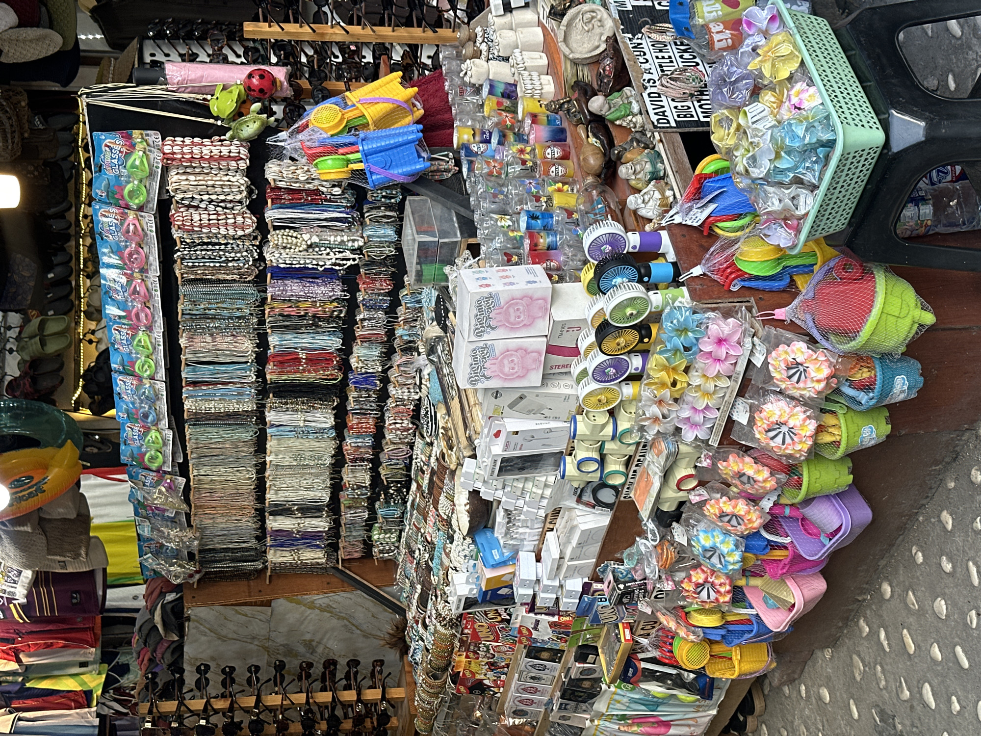 This is a photograph of a street market stall in North Kuta, Indonesia, displaying a variety of merchandise. The stall is densely packed with items arranged on shelves and hanging displays. Prominently featured are rows of necklaces and bracelets made of various beads and shells, sunglasses displayed on racks, and packaged toys such as mini fans and beach sets. Several baskets filled with colorful flower hair clips and other small accessories are also visible. In the background, clothing items and other miscellaneous goods are partially visible. The market appears to be located on a paved walkway.