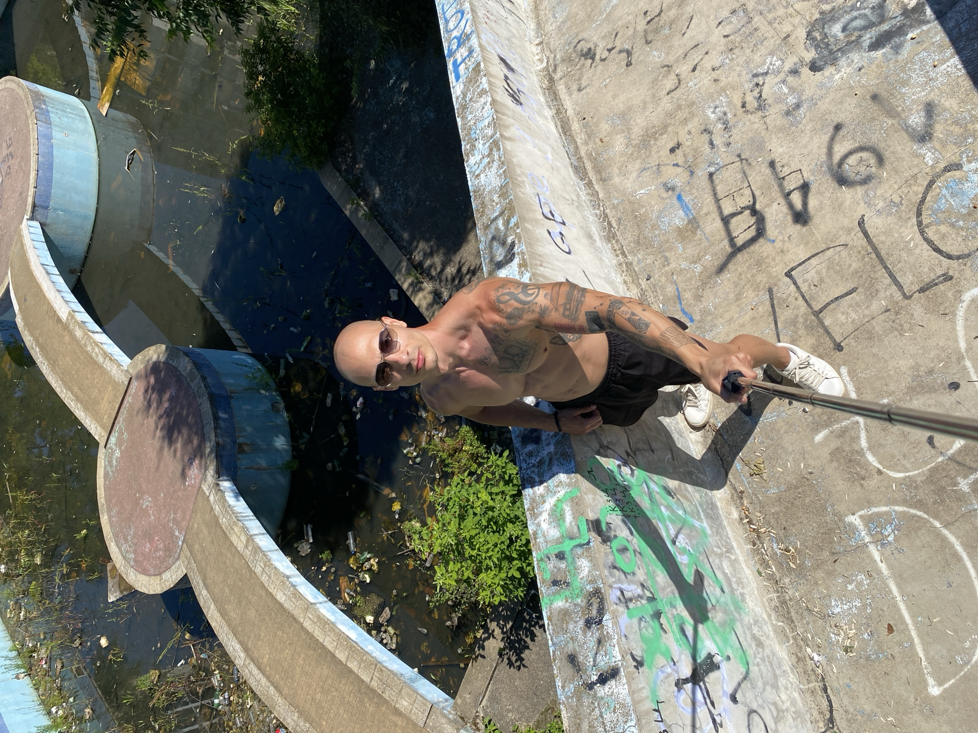 A shirtless man with tattoos on his arms is taking a selfie with a selfie stick in a dilapidated outdoor area. He is wearing sunglasses and black shorts. The background features a murky, debris-filled pool or fountain with overgrown vegetation. Graffiti covers the concrete walls surrounding the area. The scene appears to be in an abandoned or neglected public space. This location is in Pleven, Bulgaria, and appears to be an abandoned swimming pool.