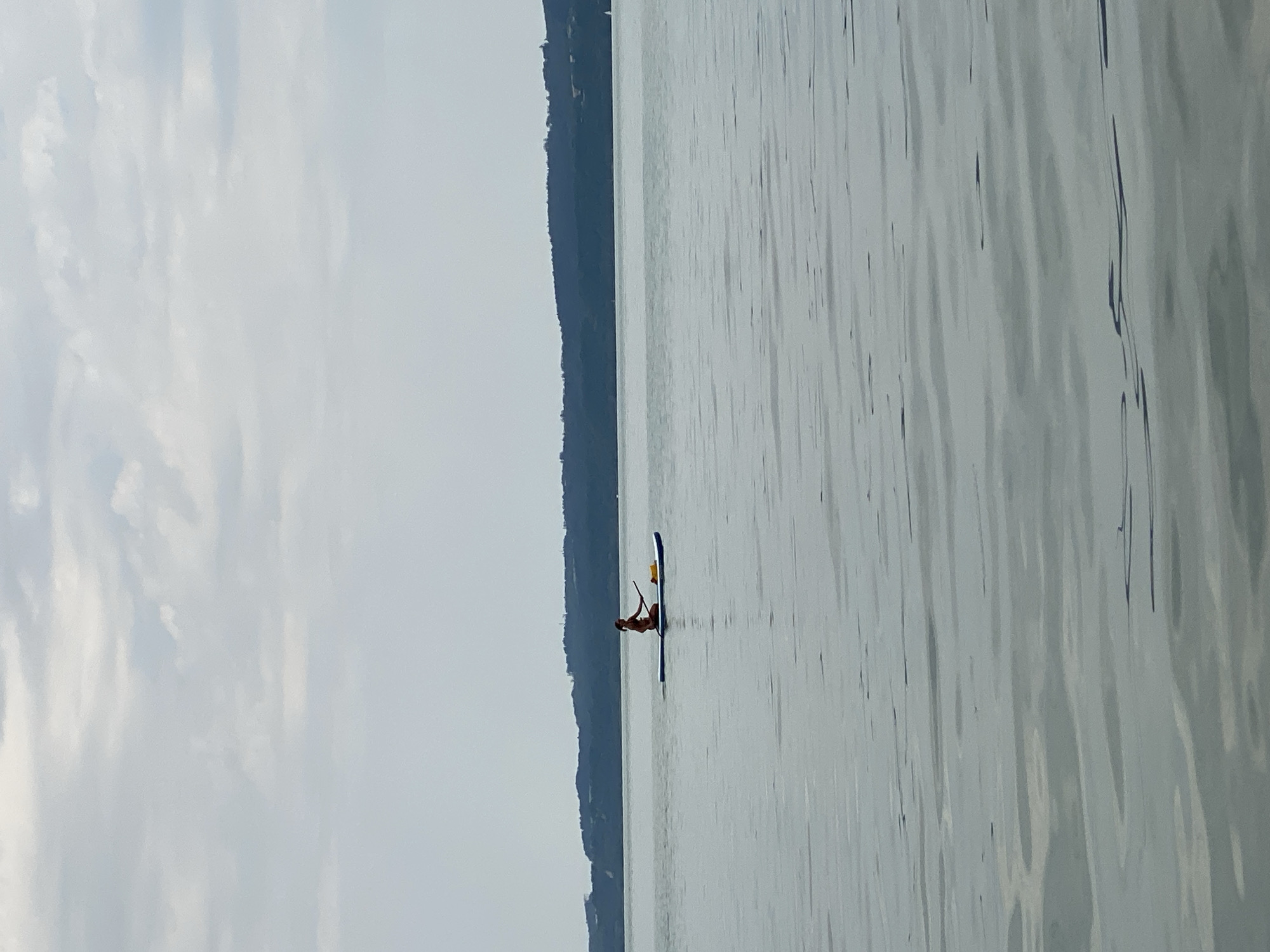A person is paddleboarding on a lake with a tree-covered hill in the background. The sky is overcast with clouds. The water is calm and reflects the sky. The paddleboarder is wearing a bathing suit and is holding a paddle. There is a yellow bag on the paddleboard. The scene is serene and peaceful, suggesting a quiet day on the water. The location is the Lake Neuchâtel in Cortaillod, Switzerland.
