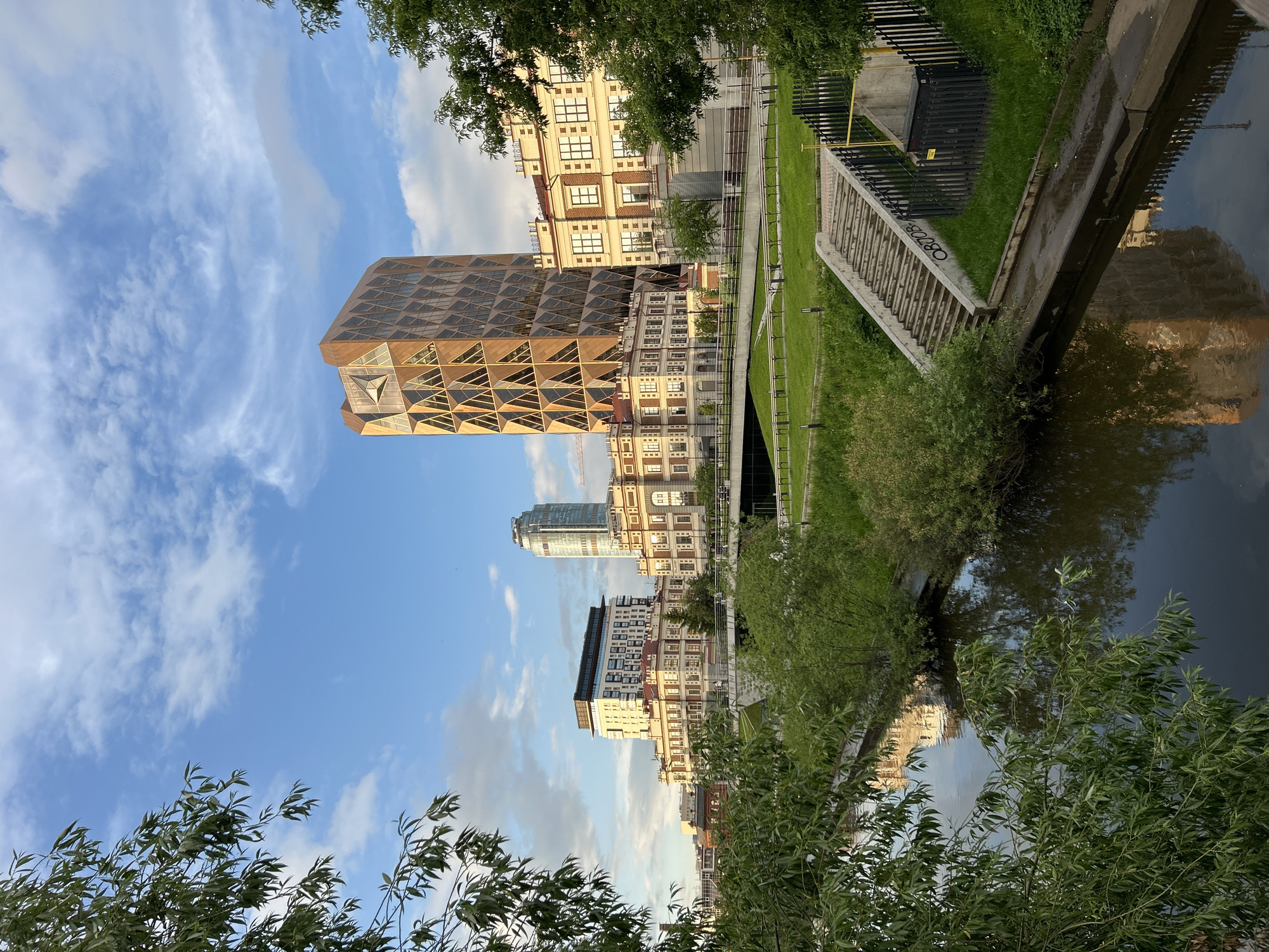 This image presents an urban landscape in Yekaterinburg, Russia, under a blue sky with scattered white clouds, illuminated by warm, low-angle sunlight indicative of late afternoon or early evening.
A narrow river with dark, reflective water occupies the lower portion of the frame. Green foliage, primarily from a deciduous tree, partially obscures the left foreground, with its branches extending over the water. Both riverbanks are lined with green grass and various bushes.
On the right bank, a concrete staircase, comprising approximately 20 steps, descends towards the water. A black metal fence borders the right side of the staircase, behind which a black utility box is visible.
In the midground and background, a cityscape is present. A prominent, tall skyscraper with a golden-brown, geometrically patterned facade featuring triangular window units stands centrally. A large, multi-pointed, star-shaped element is integrated into its upper section. Adjacent to and surrounding this skyscraper are several multi-story buildings with light-colored facades, dark roofs, and numerous windows, some displaying classical architectural features. Further in the background, to the left, a tall, slender glass skyscraper is partially visible. A multi-level bridge or elevated pathway crosses the river in the midground, with small, indistinct human figures discernible on its upper surface. Landscaped green areas and paved walkways are visible behind the riverbank structures. Reflections of the buildings are clearly visible in the calm river water.