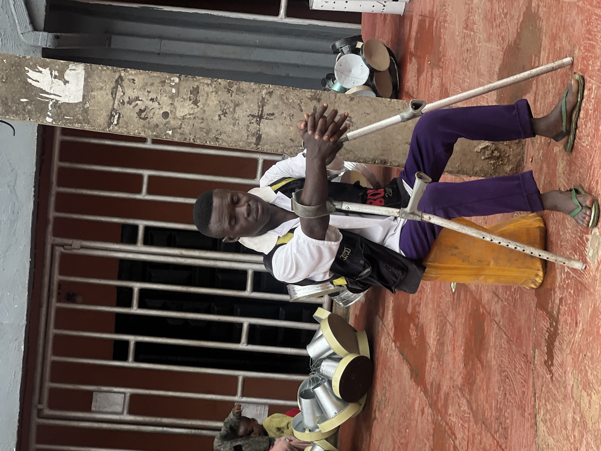 A man with crutches sits on a yellow plastic container on a red tiled surface in Nkpor, Nigeria. He is wearing a white short-sleeved shirt with a yellow and black collar, a black vest with a yellow trim, and purple trousers. His hands are clasped in his lap, and he is looking down with his eyes closed. Two crutches are positioned in front of him, supporting his arms. To his left, a group of metal containers with yellow rims are stacked on the ground. To his right, more similar containers are visible. In the background, a building with orange walls and a metal grate-covered door is partially visible. A young child is partially visible to the left of the man. The overall scene appears to be outdoors, likely in a street or marketplace.