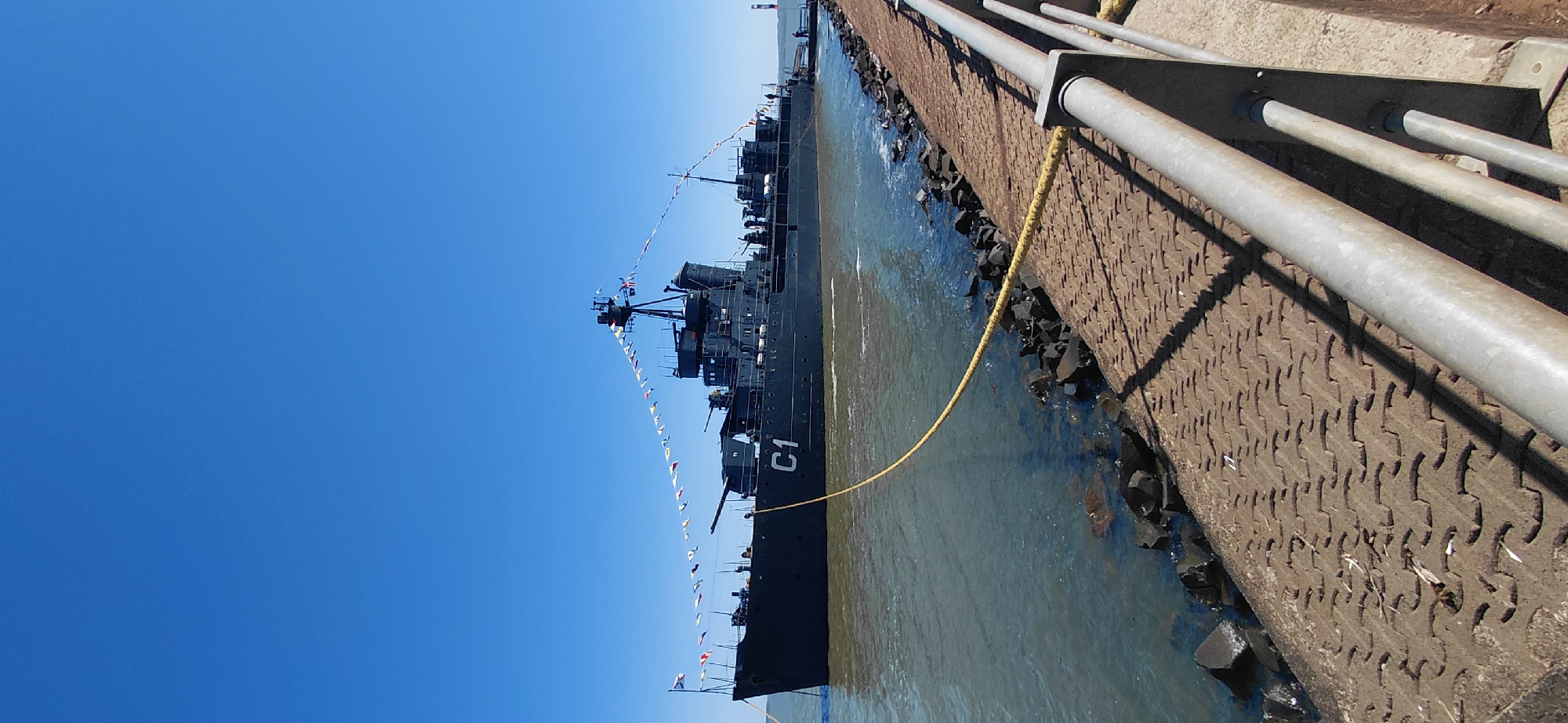 The image shows a historical navy destroyer, the "C1", docked at a pier in Encarnacion, Paraguay. The ship is dark gray with several visible gun turrets and is adorned with colorful signal flags strung along its length. A bright yellow rope is secured to the ship and extends to the pier. The pier itself is made of textured concrete and is bordered by a metal railing. Rocky formations are visible along the water's edge. The water is a murky green, and the sky above is a clear, bright blue. There are no people clearly visible on the ship, but a few figures are present on the pier in the distance. The overall scene suggests a sunny day and a static display of a naval vessel.