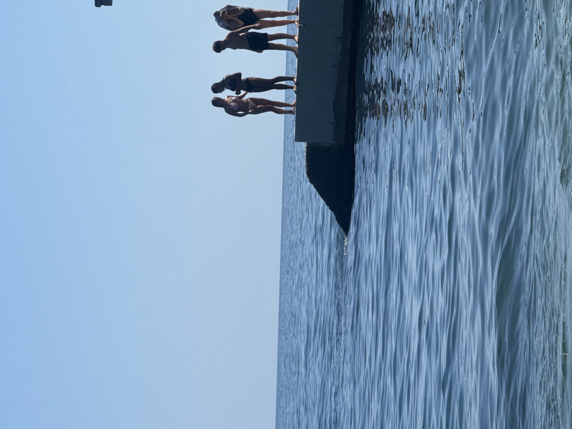 Four individuals, two females and two males, stand on a concrete pier at the edge of a body of water. The females are wearing bikinis, one in pink and the other in black. One male is wearing black shorts and the other is wearing black shorts and has a towel draped over his head. They appear to be interacting with each other, possibly conversing. The water is calm and blue, with gentle ripples on the surface. The sky is clear and blue, indicating a sunny day. The pier is grey and weathered, suggesting it has been in place for some time. The overall scene depicts a leisurely summer day at the beach. The location is the sea, near Utios, Ukraine.