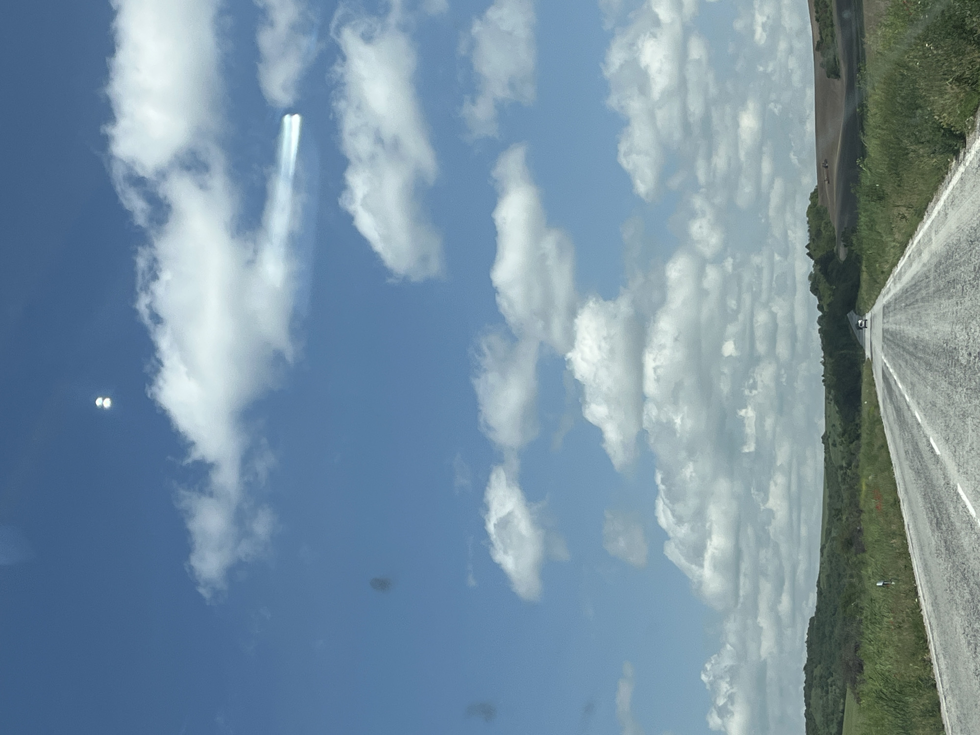 The image shows a view from a car, looking up at the sky and to the right at a road and adjacent grassy area. The sky is mostly blue with scattered white clouds. One area in the sky has a distinct, bright contrail-like formation. To the right, a straight, grey asphalt road stretches into the distance, flanked by green vegetation. A car is visible on the road in the distance. The image was taken on a roadside, at a location near Karaisen, Bulgaria.