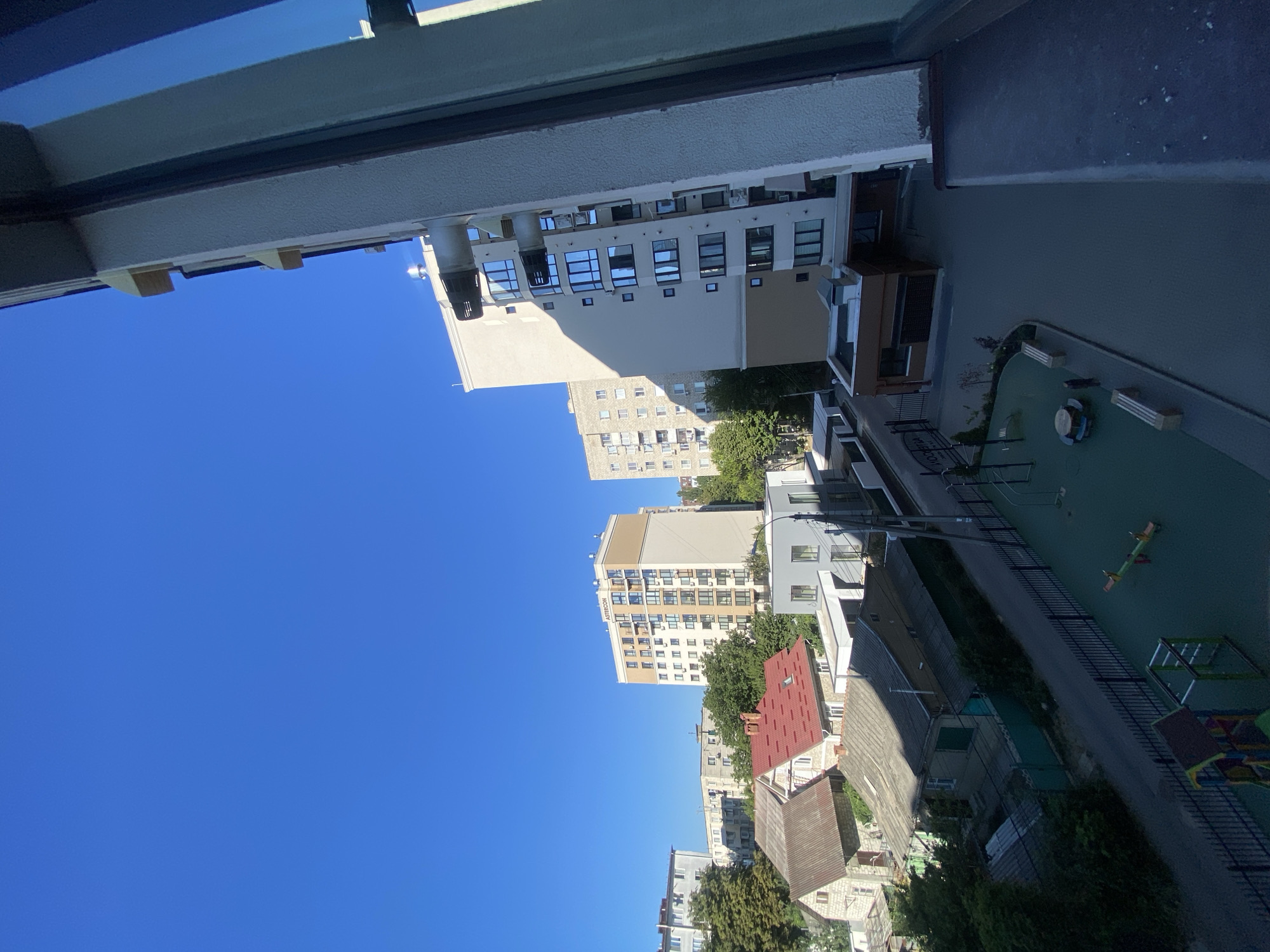 This is a high-angle, eye-level shot of a city neighborhood on a clear, sunny day. The sky is a vivid, cloudless blue. In the foreground, the edge of a modern building, possibly an apartment complex, is visible on the right side, with a dark window and a textured, off-white exterior wall. Below this building, a courtyard area with a playground is visible. The playground includes a seesaw, a small play structure with colorful elements, and two benches.
Beyond the courtyard, a street runs through the neighborhood, lined with a variety of buildings. To the left of the street, several older, smaller houses with red and gray roofs are nestled amongst trees. Further in the background, several multi-story apartment buildings of varying architectural styles are visible, some with light-colored facades and numerous windows. A sign with Cyrillic lettering can be seen on one of the apartment buildings. The overall impression is of a developing urban area with a mix of residential structures. This scene is consistent with many neighborhoods in Chișinău, Moldova, particularly areas that have seen recent construction alongside older housing.