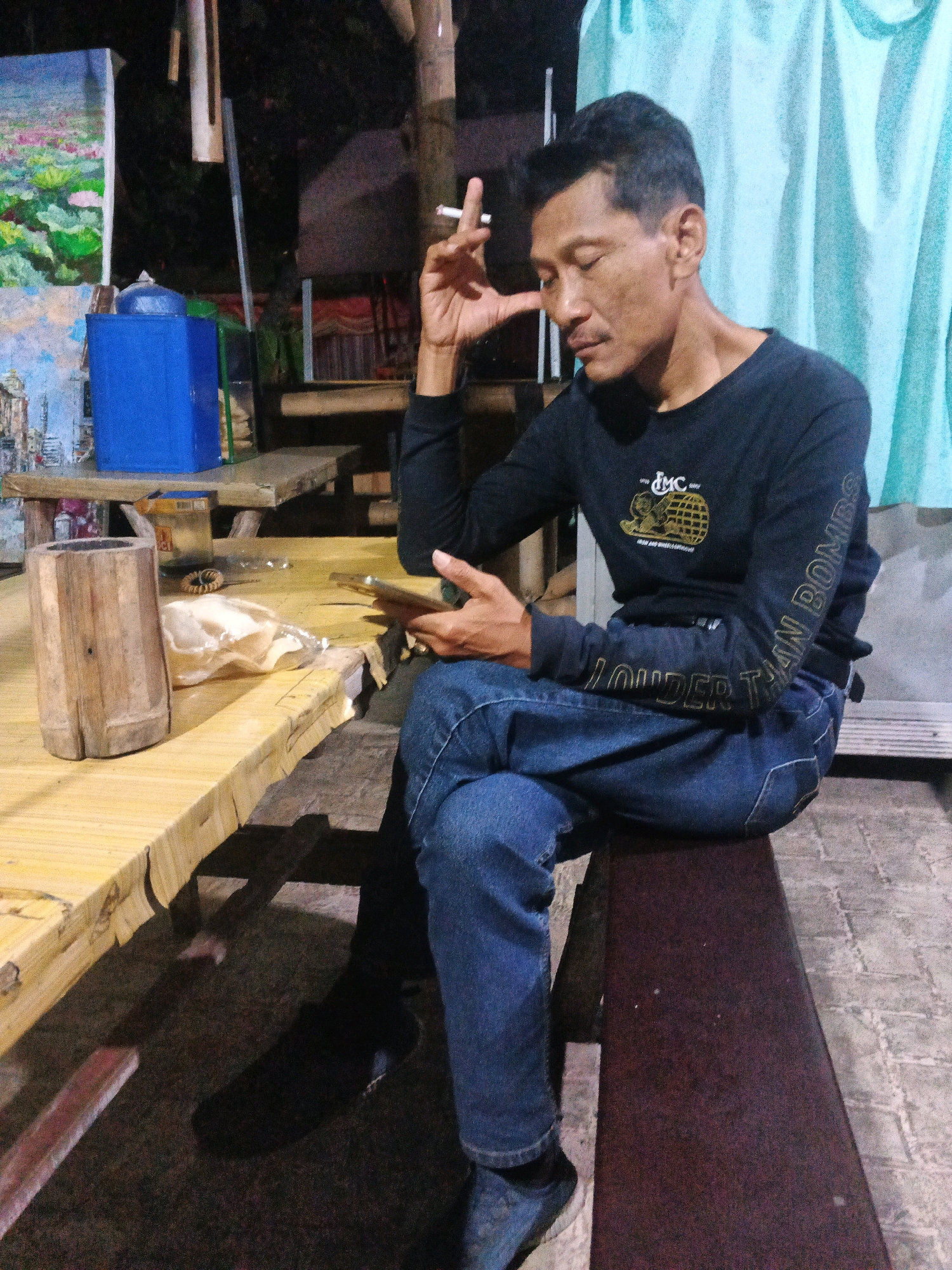 A man is sitting at a wooden table outdoors at night, smoking a cigarette and looking at a mobile phone. He is wearing a dark long-sleeved shirt and blue jeans. The table is made of light-colored wood and has some items on it, including a cylindrical wooden object, a plastic bag with food, and a coil of wire. Behind the man is a light blue curtain. In the background, there are paintings and structures, possibly indicating a local market or street stall. The lighting suggests it is evening or night. The location is identified as Ungaran, Indonesia.