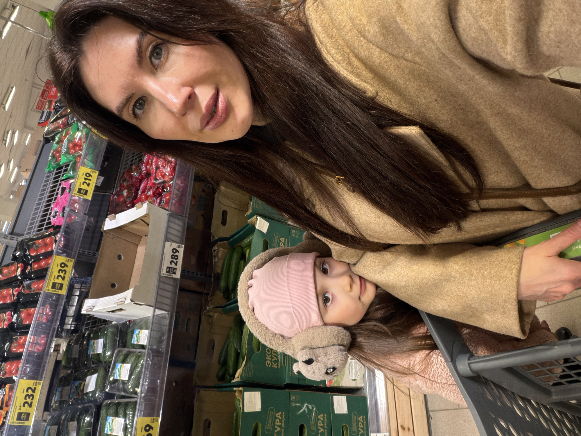 The image captures a selfie-style view of a woman and a child in what appears to be a grocery store. The woman, presumably the mother, is in the foreground with dark brown hair and is wearing a tan-colored coat. Her face is clearly visible, and she seems to be looking at the camera. Next to her, a young child, presumably her daughter, is wearing a pink beanie-style hat with fuzzy earmuffs resembling animal ears. She is sitting in a shopping cart.
The background reveals the interior of a grocery store aisle. Shelves stocked with produce, specifically tomatoes and cucumbers, are visible. Price tags are present on the shelves, displaying prices like "232," "239," and "289," suggesting a market setting. Based on the visible text "КУРА" and "УРА", as well as the Russian denominations on the price tags, this grocery store is located in Russia, more specifically the city of Perm.
