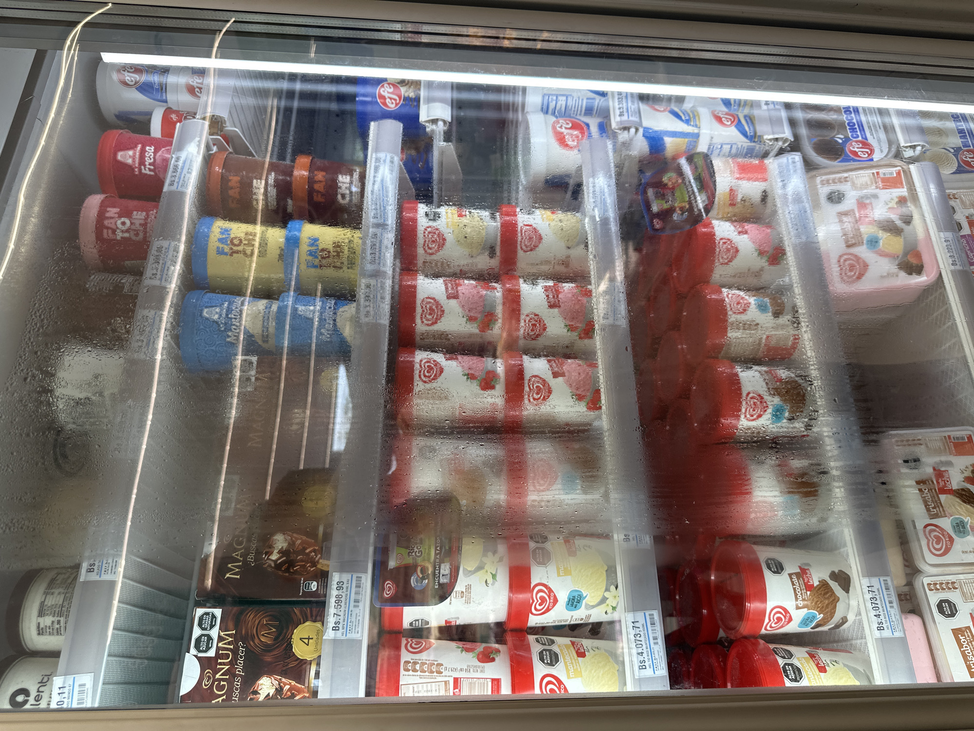 The image shows the inside of a commercial freezer display case, likely in a convenience store or supermarket. The interior of the freezer is visible through a glass door covered in condensation. Various ice cream products are displayed on shelves.
On the top shelf, there are large tubs of ice cream with labels like "FAN TO CHE," "LA ARGENTINA Fresa," and "efe." There are also some boxes of "MAGNUM" ice cream bars.
The middle shelf contains rows of smaller tubs of ice cream. Brands include "TIO RICO" with flavors like "manleado" (likely "manjar" or dulce de leche), and other tubs with images of strawberries and chocolate.
The bottom shelves also display ice cream tubs and some multipacks of ice cream bars with similar branding to those on the middle shelf. Some of the tubs have clear plastic lids.
The labels on the price tags are in Bolivars (Bs.), indicating a Venezuelan currency. One price tag is visible for "HEL MPK MAGNUM ALMENDRA 90ML X 4U." Another price tag visible on a different shelf reads "Bs.4.073,71."
The overall environment is cold and humid, as evidenced by the condensation on the glass. The lighting appears to be from within the display case, casting a cool glow on the products. There are no people visible in the image.