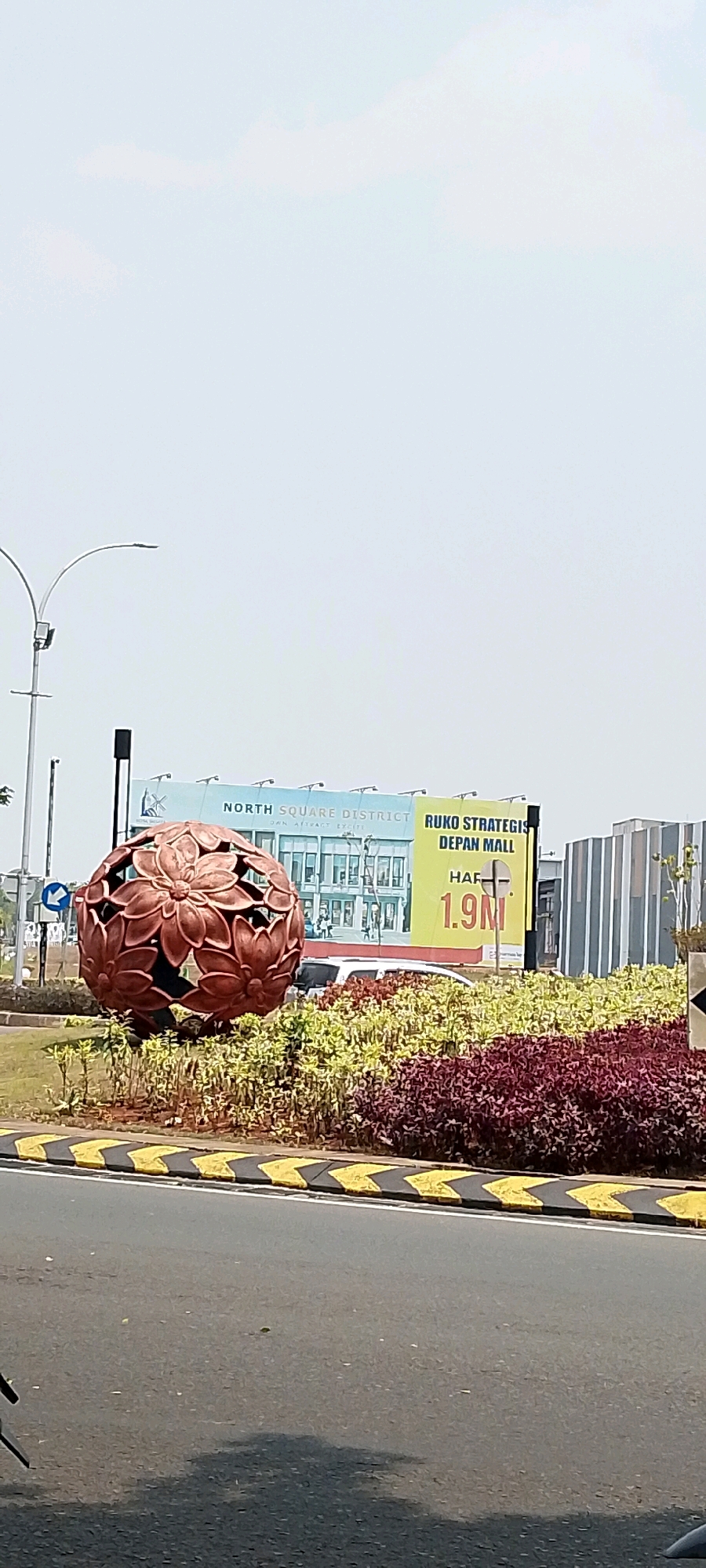 The image depicts an outdoor scene in the city of Gunung Putri, Indonesia, characterized by a large, ornate, copper-colored spherical sculpture resembling a flower or lotus, positioned prominently in a landscaped area. The sculpture is surrounded by a variety of green and reddish-purple plants, set against a backdrop of a road with yellow and black chevron markings. Behind the sculpture and landscaping, a billboard is visible. The billboard features text in Indonesian, advertising "NORTH SQUARE DISTRICT" and "RUKO STRATEGIS DEPAN MALL HARGA 1.9M", along with an image of a building and a mall. Further in the background, there are modern-looking buildings, one with a distinctive striped facade. A streetlamp with two curved arms is also present to the left of the scene. The sky is overcast with a light haze. No people are visible in the image. The overall impression is one of a well-maintained urban public space, likely near a commercial or residential development.