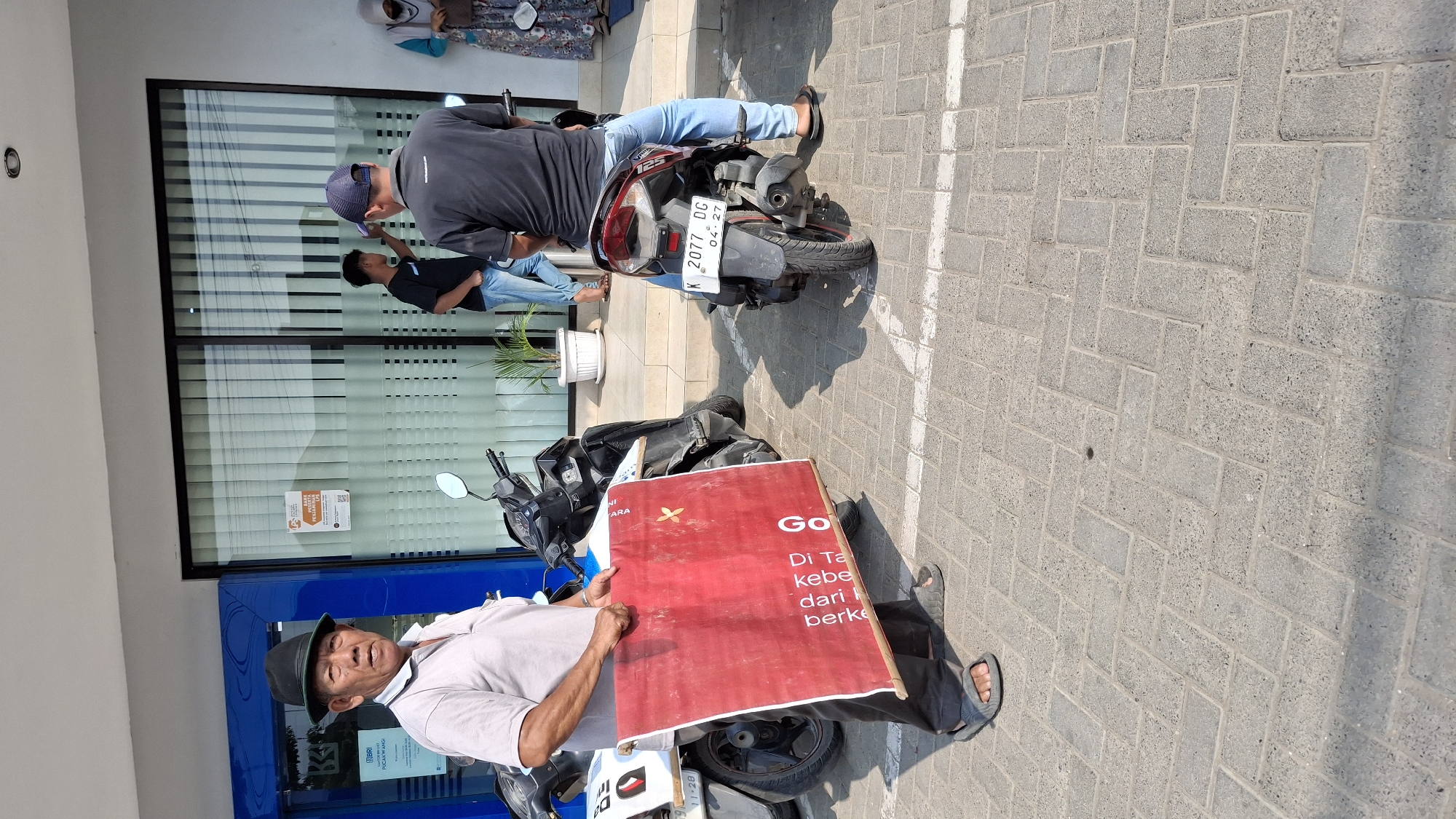 An outdoor daytime medium shot shows an elderly Indonesian man wearing a grey polo shirt, black pants, grey sandals, and a black fedora hat. He is standing on a paved surface and holding a large, red banner with some text on it. The banner appears to be made of fabric and is held taut by wooden poles. Behind him, a black motorcycle is parked.
Further back, two younger men are visible. One, wearing a black t-shirt and blue jeans, stands near a glass-fronted building with vertical blinds. The other, wearing a dark blue t-shirt, blue jeans, and a blue baseball cap, sits on a dark-colored motorcycle that has a license plate with the inscription "K 2077 DG 04-27". A woman in a floral patterned dress and headscarf is also seen in the background, near the entrance of the building. The building appears to be a commercial establishment, possibly a bank, given the blue signage that resembles the BRI bank logo. The ground is paved with interlocking grey bricks, and white lines mark parking spaces. The location is identified as Pucakwangi, Indonesia.