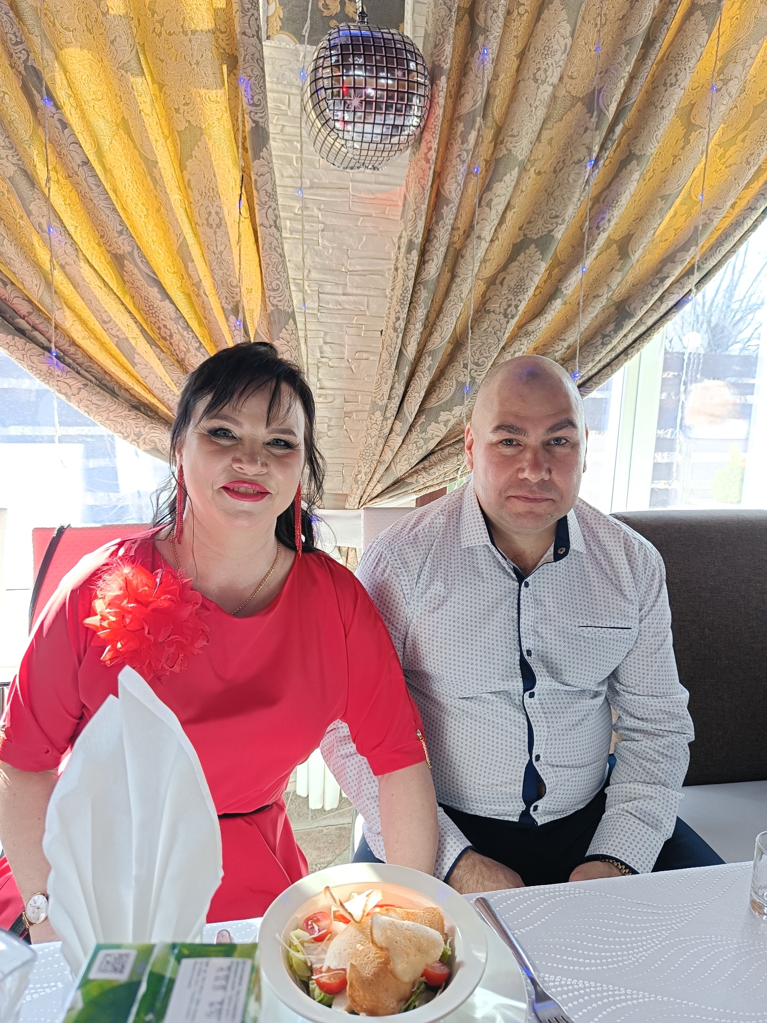 This full-shot image captures a man and a woman seated at a table in what appears to be a restaurant or banquet hall in Chaikovsky, Russia. Both individuals are looking directly at the camera.
The woman, positioned on the left, is smiling and wears a vibrant red long-sleeved dress, featuring a large red fabric flower on her left shoulder/chest. She has dark hair, bright red lipstick, and long red dangling earrings, complemented by a gold necklace and a watch on her left wrist. The man, seated on the right, is bald and wears a white long-sleeved collared shirt with a subtle dark pattern and dark pants. He has a neutral expression.
They are seated at a table covered with a white tablecloth featuring a textured pattern. Visible on the table are a neatly folded white napkin, a white bowl containing a salad with lettuce, cherry tomatoes, and croutons, a fork, and a clear glass. A green box or menu item with a visible QR code is partially seen on the table.
The background features heavy, ornate curtains in yellow/gold and grey/beige patterns, draped decoratively. Small blue LED fairy lights are strung along these curtains, creating a festive ambiance. A silver disco ball hangs from the ceiling. Bright natural light streams through large windows behind them, indicating a sunny daytime setting. Blurry outlines of outdoor trees or buildings are visible through the windows. The woman is seated on a red chair, while the man is on a dark brown booth-style seat. The scene suggests a pleasant dining experience or celebration.