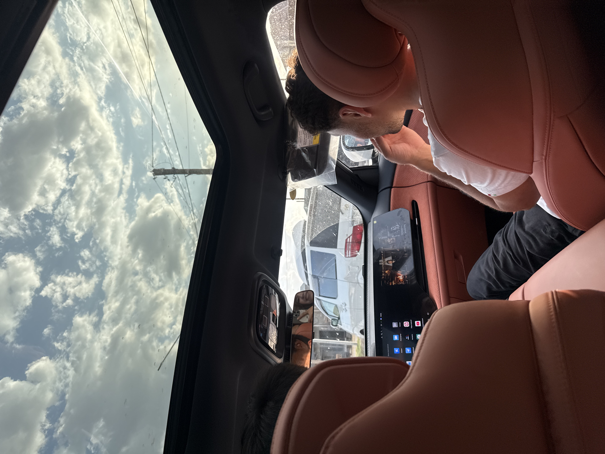 The image shows the interior of a car, looking towards the front. The car has a panoramic sunroof, through which a cloudy sky is visible. In the foreground, the back of two car seats with orange leather upholstery are visible. In the middle ground, a man with dark curly hair is sitting in the passenger seat, looking to his right and touching his chin with his hand. He is wearing a white t-shirt. The car's dashboard displays a touchscreen interface with various icons and a video playing, featuring a title that reads "TECHNO MAFIA." The driver's side rearview mirror reflects the faces of two people in the back seat, one holding a smartphone. Outside the car, through the windows and sunroof, a white van and a utility pole with wires are visible. The location is identified as Şardara, Kazakhstan.