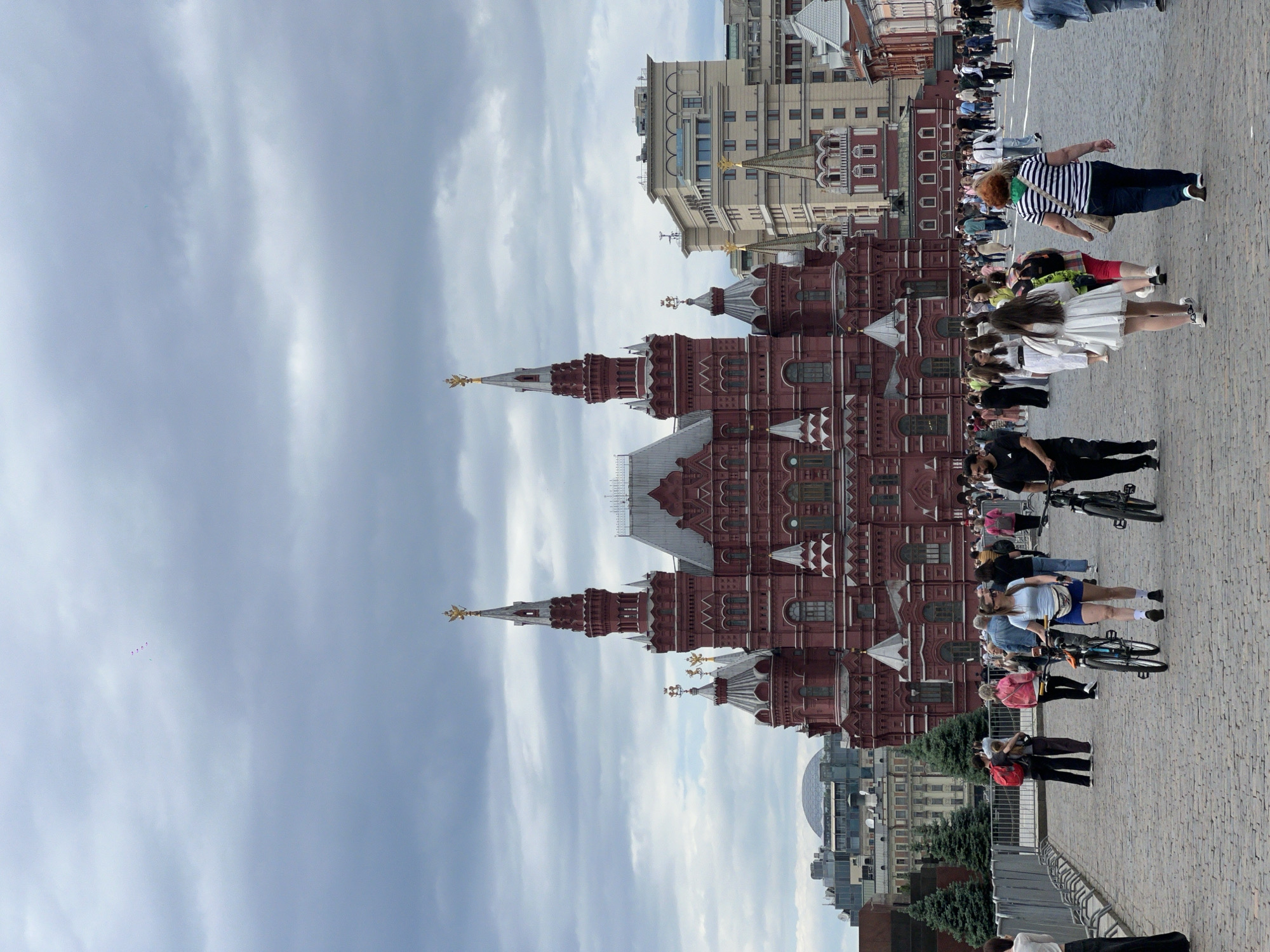 Here is a description of the image:
The image shows a view of the State Historical Museum in Moscow, Russia, a large red building with pointed towers. The museum is surrounded by a plaza made of gray cobblestones. There are many people walking and riding bikes in the plaza. The sky is cloudy and gray. Other buildings are visible in the background.