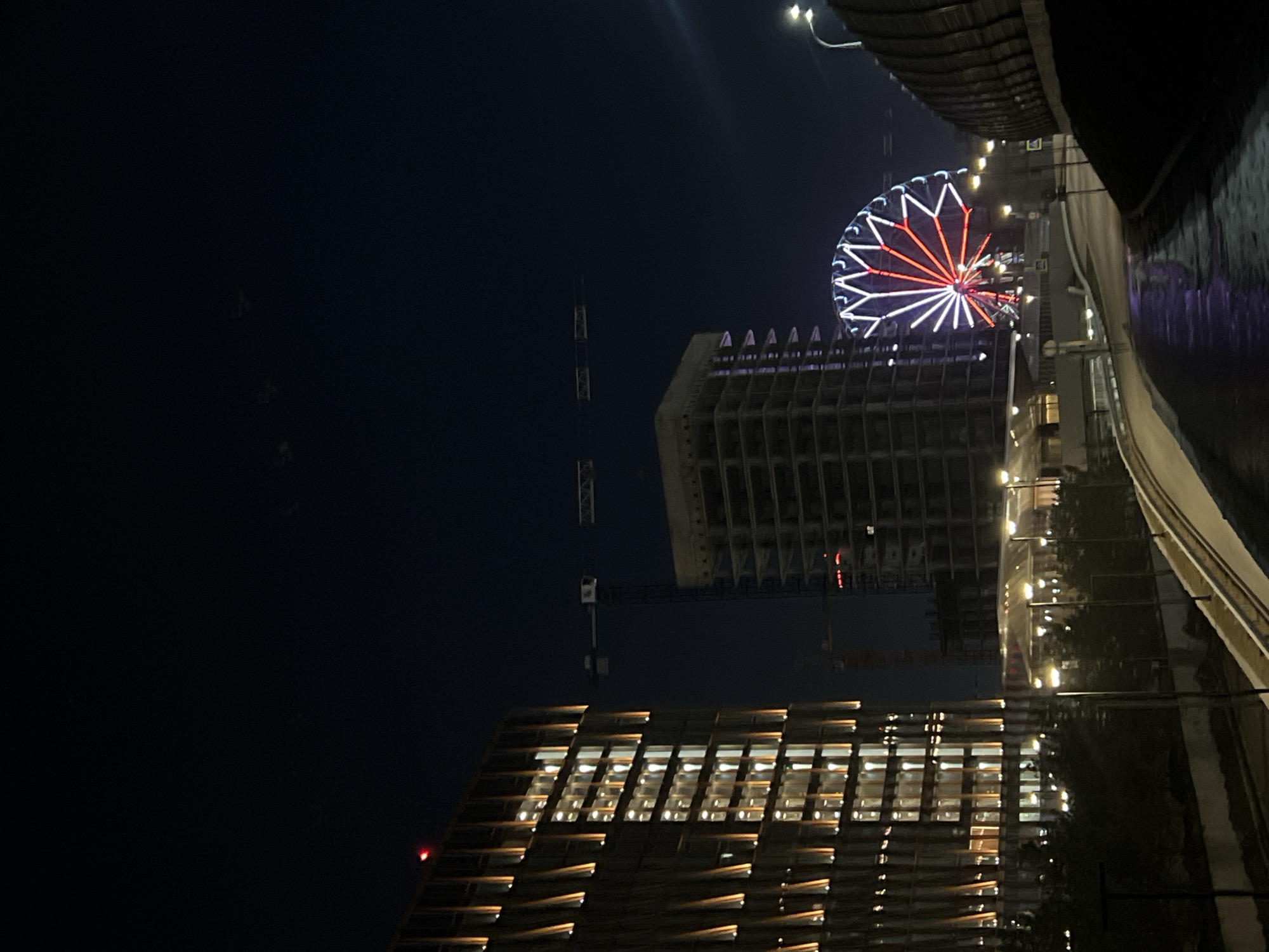 At night, a building with lights visible through its windows stands out prominently in the foreground. Above it, a Ferris wheel with red and white lights is illuminated against the dark sky. A building under construction is also visible. The scene appears to be located near a body of water reflecting the city lights. The Kazan Riviera is visible to the right, indicating that this could be somewhere in the city of Kazan, Russia.