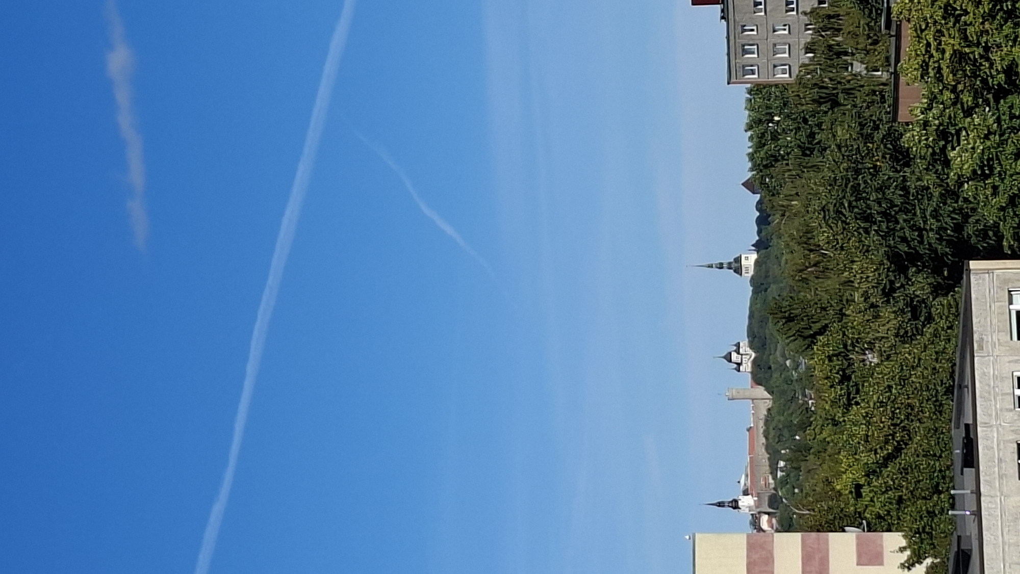 The image captures a view of Tallinn, Estonia, on a clear day with a bright blue sky. Two distinct contrails are visible stretching across the sky. In the middle ground, a cluster of buildings with distinctive architecture, likely historical, is situated on a hill covered with lush green trees. These buildings include spires and towers, suggesting a religious or governmental complex. To the left and right of this central scene, modern buildings are partially visible. On the far left, a section of a building with horizontal beige and red-brown stripes is prominent. On the right, a grey building with multiple rectangular windows is seen. In the foreground, the roofs of several buildings, one appearing to be a concrete structure with visible ventilation pipes, are present. The overall impression is an urban landscape blending historical and modern elements under a vast sky.
