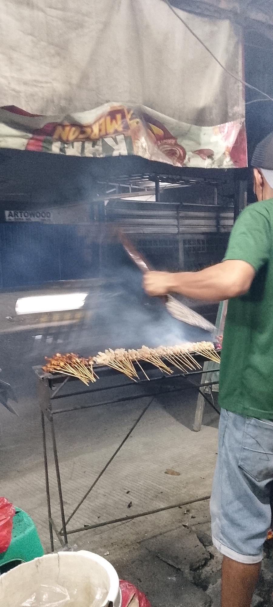 An outdoor scene in Satriamekar, Indonesia, depicts a person grilling satay. The individual, wearing a green t-shirt and denim shorts, is standing to the right of the frame, holding a tool to tend to the skewers of meat cooking on a charcoal grill. Smoke rises from the grill, obscuring parts of the background. Several rows of satay skewers are visible on the grill. In the background, there is a banner with text, part of which appears to read "ARTOWOOD." The ground is paved. To the left of the frame, there are some plastic containers and bags.