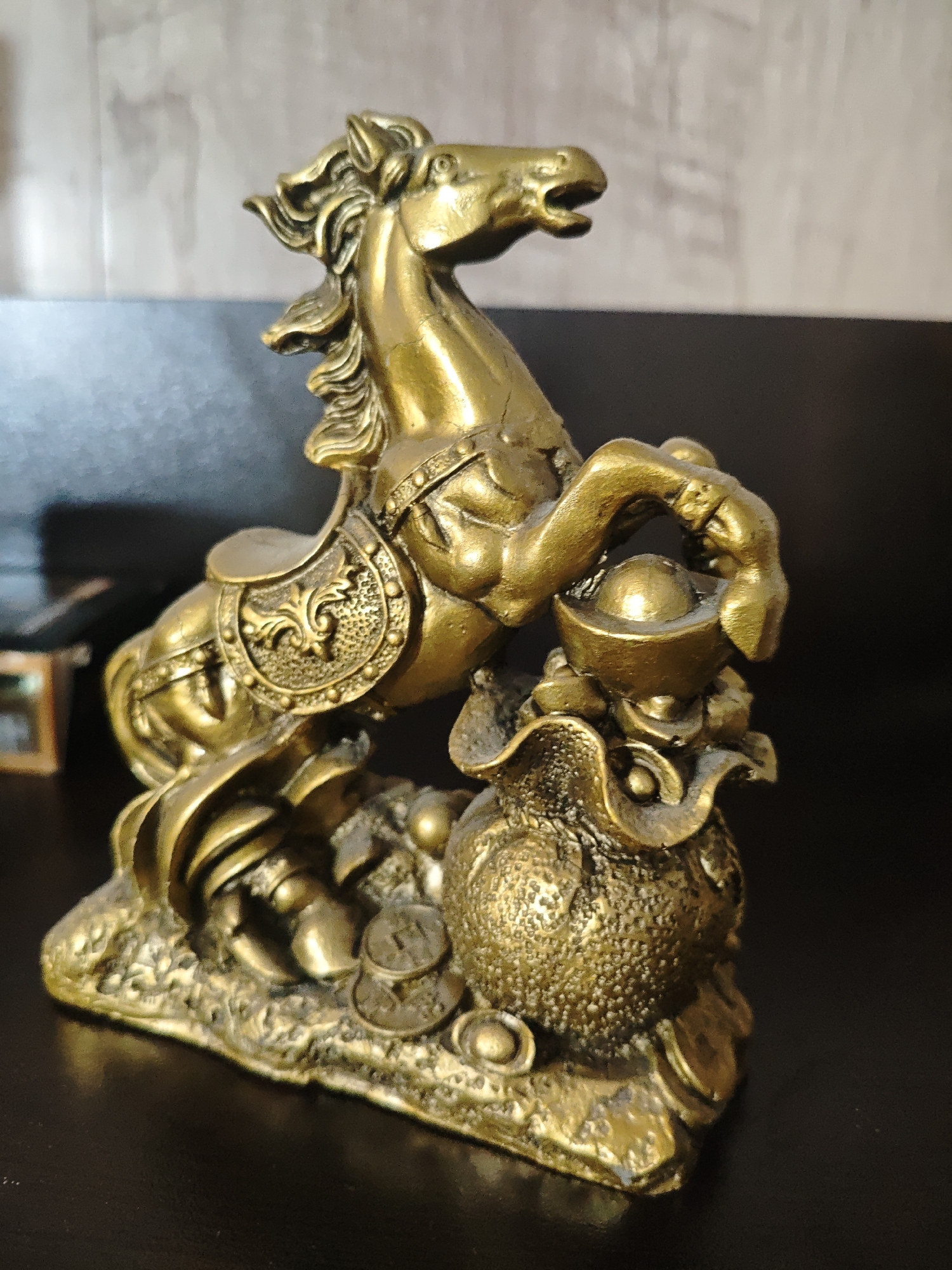A golden-colored horse statue is positioned on a dark, reflective surface. The horse is in a rearing position, with its front hooves elevated and one hoof holding a golden bowl-shaped object. The horse is elaborately detailed with ornate saddle and bridle features. Below the horse is a textured, sack-like object surrounded by several flat, coin-shaped objects. The background is blurred, consisting of a light-colored wall above a darker surface. The setting is indoors.