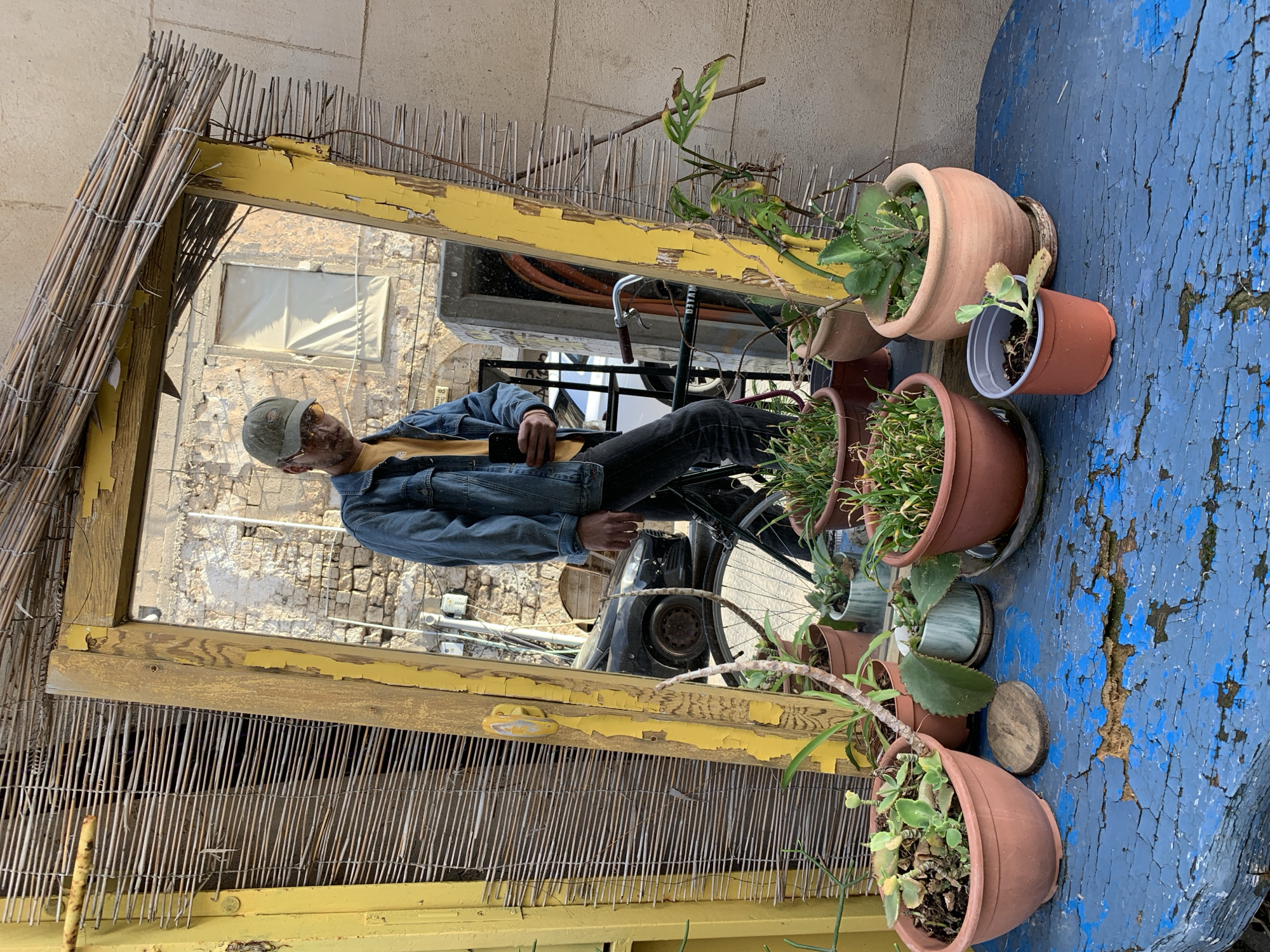 A medium shot shows a man sitting on a bicycle and reflected in a mirror, surrounded by potted plants on a distressed blue tabletop. The mirror is framed by a weathered yellow wooden structure with a thatched overhang. The man is wearing a denim jacket, a yellow shirt, dark pants, and a cap. He is looking towards the viewer. The mirror reflects a street scene with a stone building and parked cars. The tabletop features several terracotta pots filled with greenery, while the blue paint on the table is peeling and cracked. The location is Tel-Aviv, Israel, based on the architectural style and general surroundings.