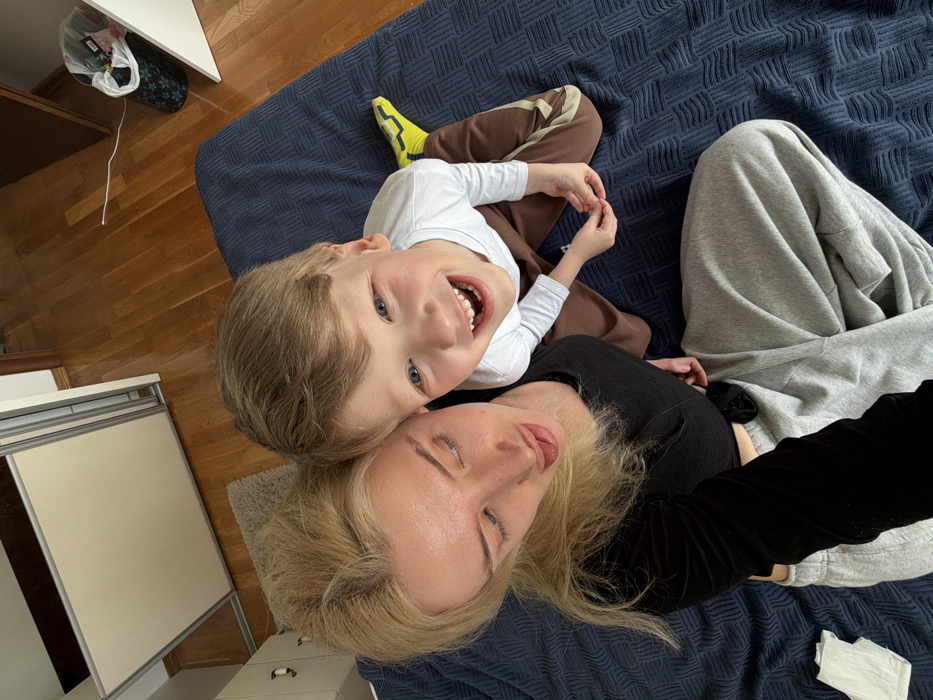 The image depicts an overhead perspective of an adult female and a young male child within an indoor residential setting. The female, positioned on the left, possesses long blonde hair and is attired in a black long-sleeved top and grey sweatpants. She gazes downwards, her mouth slightly ajar with her tongue extended, indicating a playful or candid moment while holding the camera. The child, situated to her right, features light hair and blue eyes. He presents a broad smile, looking directly upwards towards the camera. He wears a white long-sleeved shirt, brown sweatpants with light-colored stripes along the sides, and a single neon yellow and black athletic shoe on his visible left foot. The child is kneeling or seated on his knees, his hands resting together. Both individuals are positioned on a dark blue, textured blanket or duvet. To the left of the female, a crumpled white tissue is visible on the bedding. The background reveals polished hardwood flooring. A light grey, shaggy rug is partially visible on the floor, located beneath a portion of a white cabinet or dresser on the far left. A light-colored wardrobe with sliding doors is present on the left side of the frame. Towards the upper right, a dark brown wooden door frame is visible, along with a partially seen white cabinet and a black wire wastebasket featuring a floral pattern and containing some refuse. A white cable traverses the hardwood floor in the upper right quadrant. The scene is brightly and evenly lit.