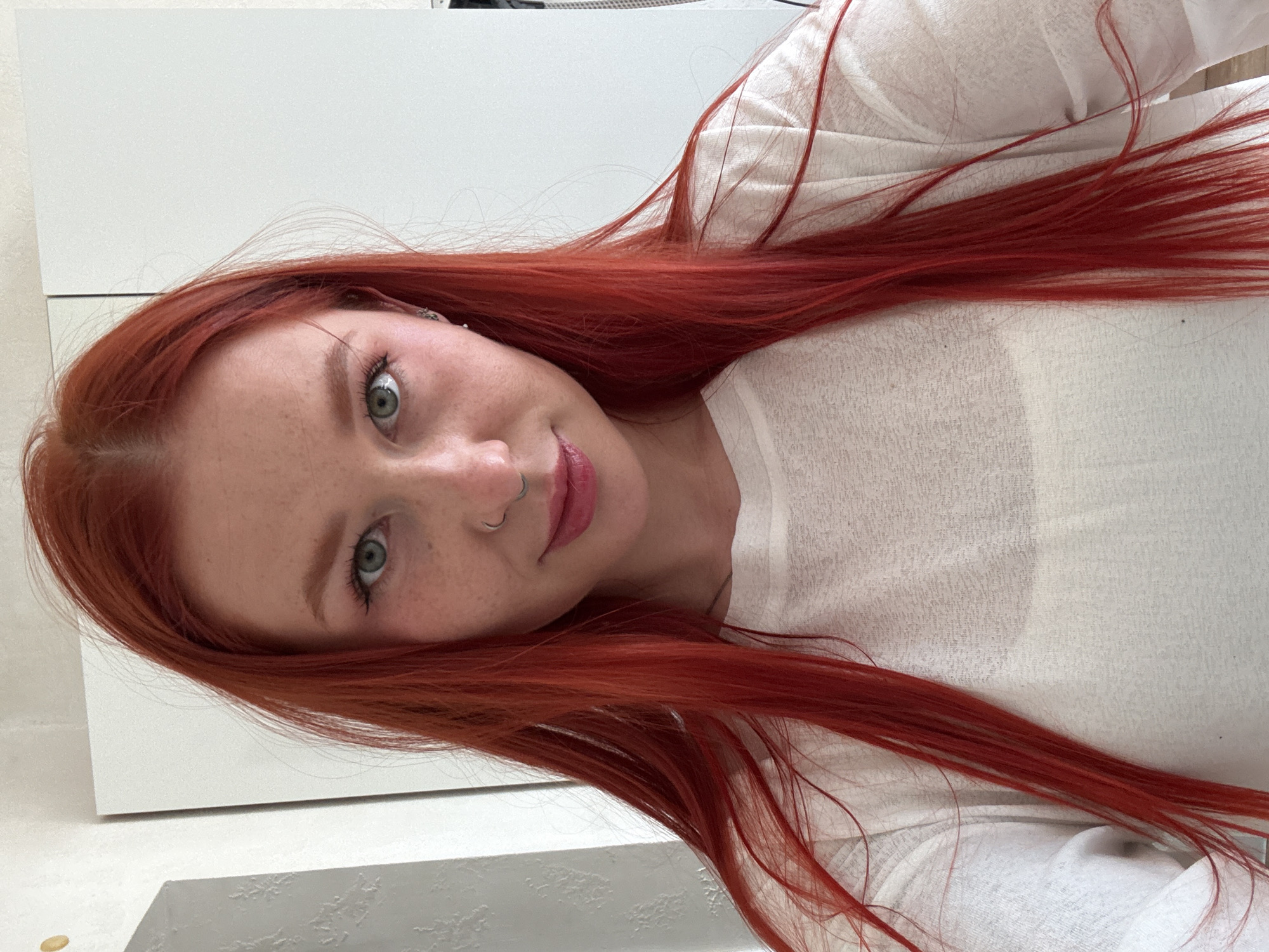 A close-up portrait shows a young woman with long, vibrant red hair and striking green eyes, looking towards the viewer with a gentle, slight smile. Her fair skin is adorned with scattered freckles, particularly visible across her nose and cheeks. She has a small silver hoop nose ring in her left nostril. Her lips are slightly parted, revealing a natural pink hue. She is wearing a light-colored, possibly white or off-white, long-sleeved top made of a soft, somewhat sheer material. The background is minimalist and bright, appearing to be an indoor setting with white walls or furniture. The lighting is soft and even, suggesting an interior daytime environment. No text is visible in the image.