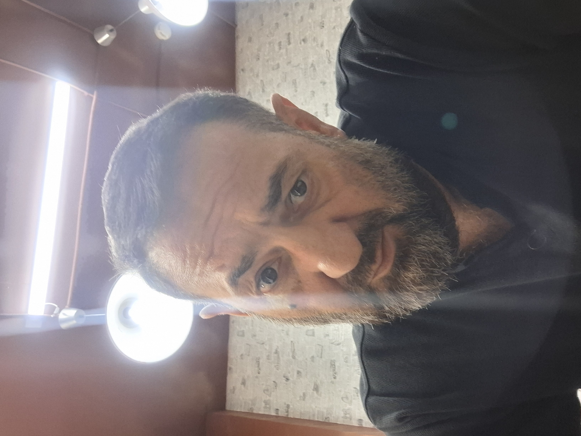 A medium shot selfie shows a man with short dark hair, a beard, and a black shirt. He is looking at the camera with his head tilted slightly to the left. The background shows a brick wall and a ceiling with lights. The man appears to be indoors, possibly in a commercial or residential setting. There are no identifiable landmarks or indications of the specific location within Moscow, Russia.