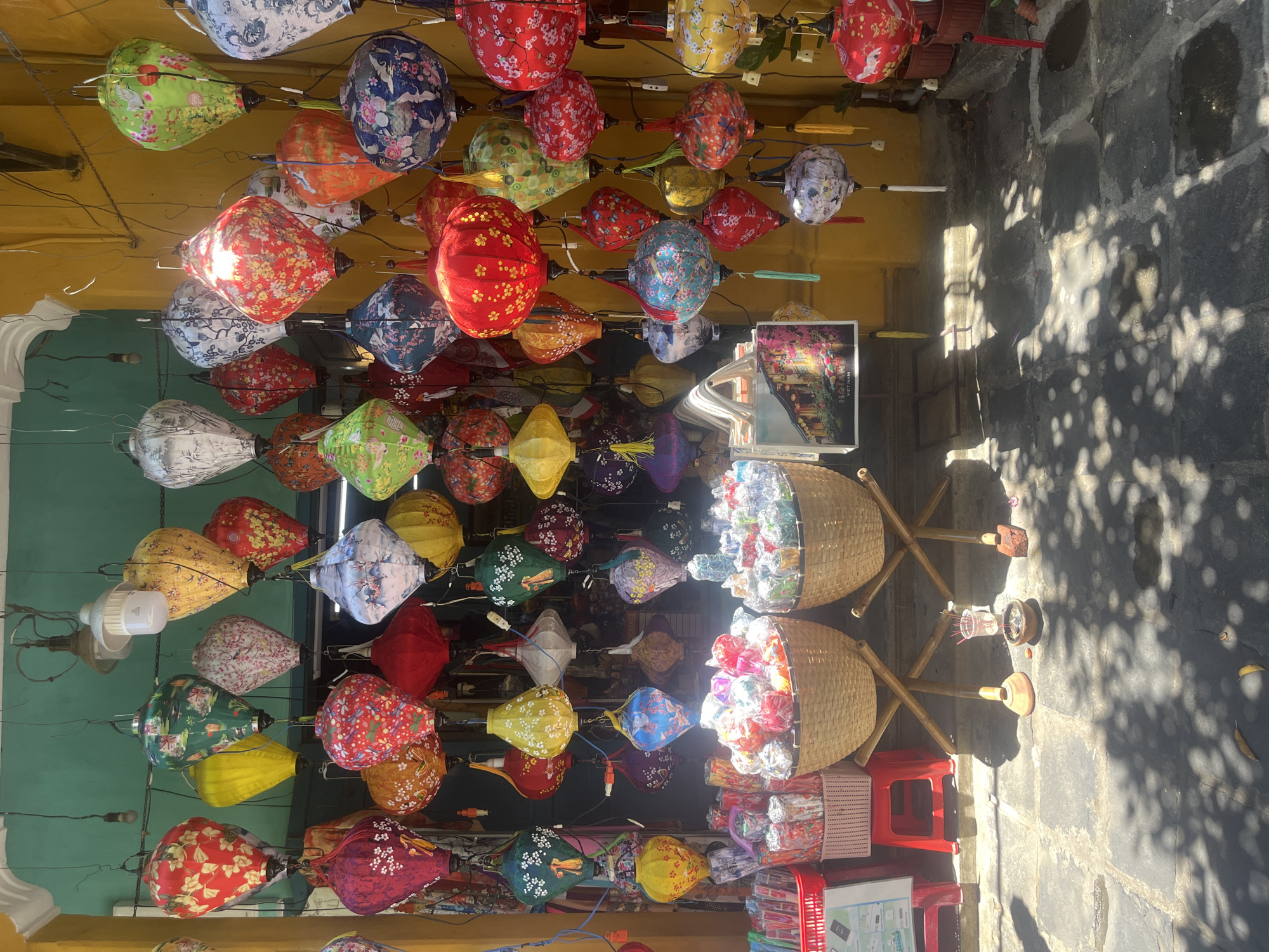 This image depicts a vibrant and colorful outdoor scene, likely a market stall or shop alleyway in Hội An, Vietnam, as suggested by the visible text and context. The dominant feature is a dense canopy of numerous traditional lanterns hanging overhead, creating an immersive and decorative display.
The lanterns are diverse in shape, including round, oval, and diamond-like forms, and feature a rich palette of colors such as red, green, blue, yellow, white, orange, and purple. Each lantern is adorned with intricate patterns, including detailed floral designs, traditional motifs, and depictions of animals like cranes and dragons. Many appear to be made of silk or a similar fabric stretched over a frame, and some are subtly illuminated, adding to their allure.
On the right side of the frame, two large, woven bamboo baskets rest on wooden stands. These baskets are filled with smaller, individually wrapped items, possibly mini lanterns or other souvenirs, displayed for sale. Below them, a red plastic stool and another smaller white basket are visible. The ground below is paved with dark stone, characterized by strong dappled sunlight and shadows, indicating a bright daytime setting. Above the lanterns, a yellow ceiling or awning is present, with a teal-green wall to the left.
A small framed picture or postcard, placed on a stand, is visible on the right, showcasing a night scene illuminated by lanterns. Clearly legible text on this item reads: "HOI AN VIETNAM." The scene conveys a lively, traditional, and visually rich atmosphere, characteristic of the area known for its lantern craftsmanship.