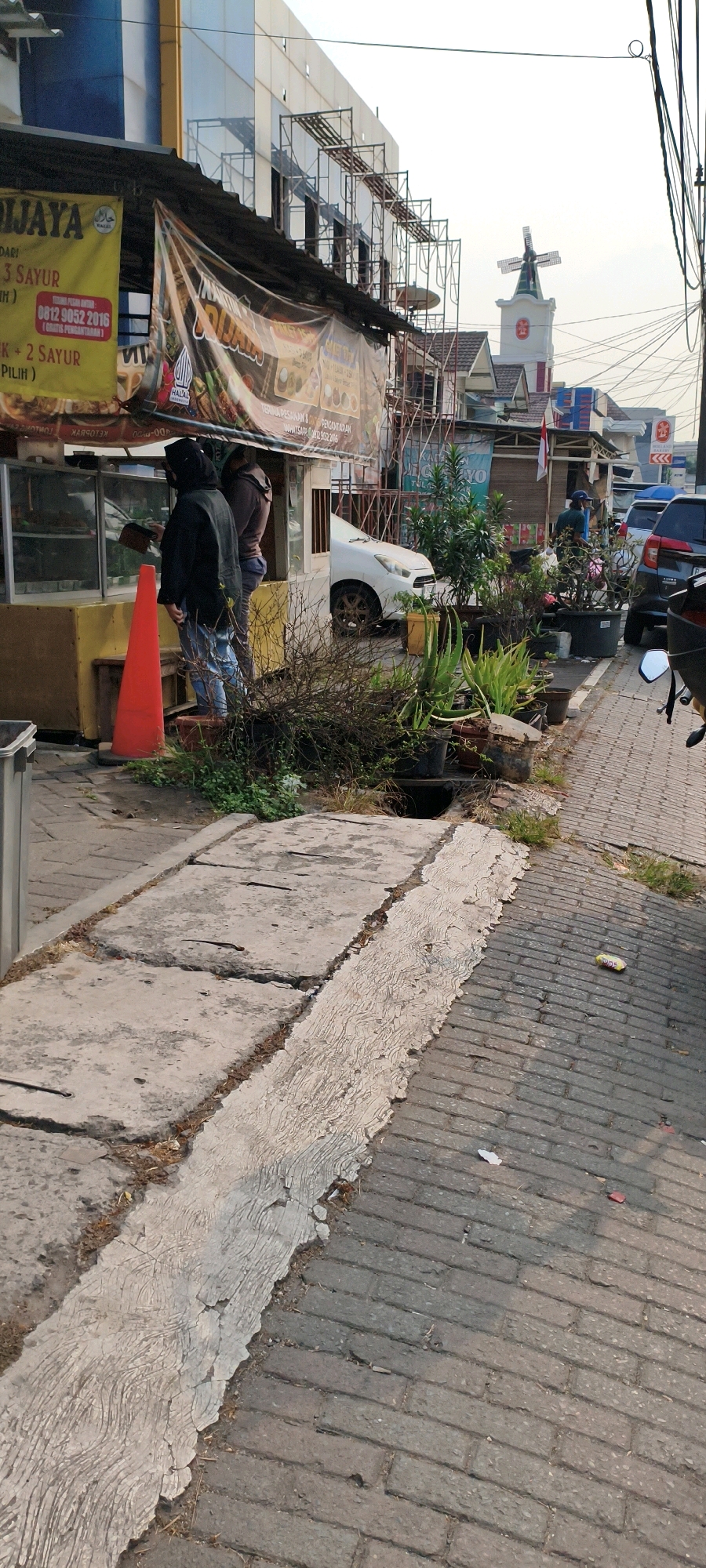 The image depicts a street scene in West Jakarta, Indonesia, focusing on a food stall area and a nearby sidewalk. In the foreground, a cracked concrete sidewalk transitions to a brick-paved road. A prominent red traffic cone stands on the sidewalk next to a food stall. Two individuals are partially visible behind the counter of the food stall. The stall itself has a canopy and banners with text, some of which appear to be advertisements.
Further back, the scene reveals scaffolding attached to a building, suggesting ongoing construction or renovation. Across the street, a white car is parked. A cluster of potted plants, including aloe vera, is arranged along the edge of the sidewalk. In the background, a building with a distinctive tower topped by a windmill is visible. Various other buildings, some with signage, and other vehicles, including motorcycles and cars, populate the streetscape. The overall lighting suggests it is either early morning or late afternoon.