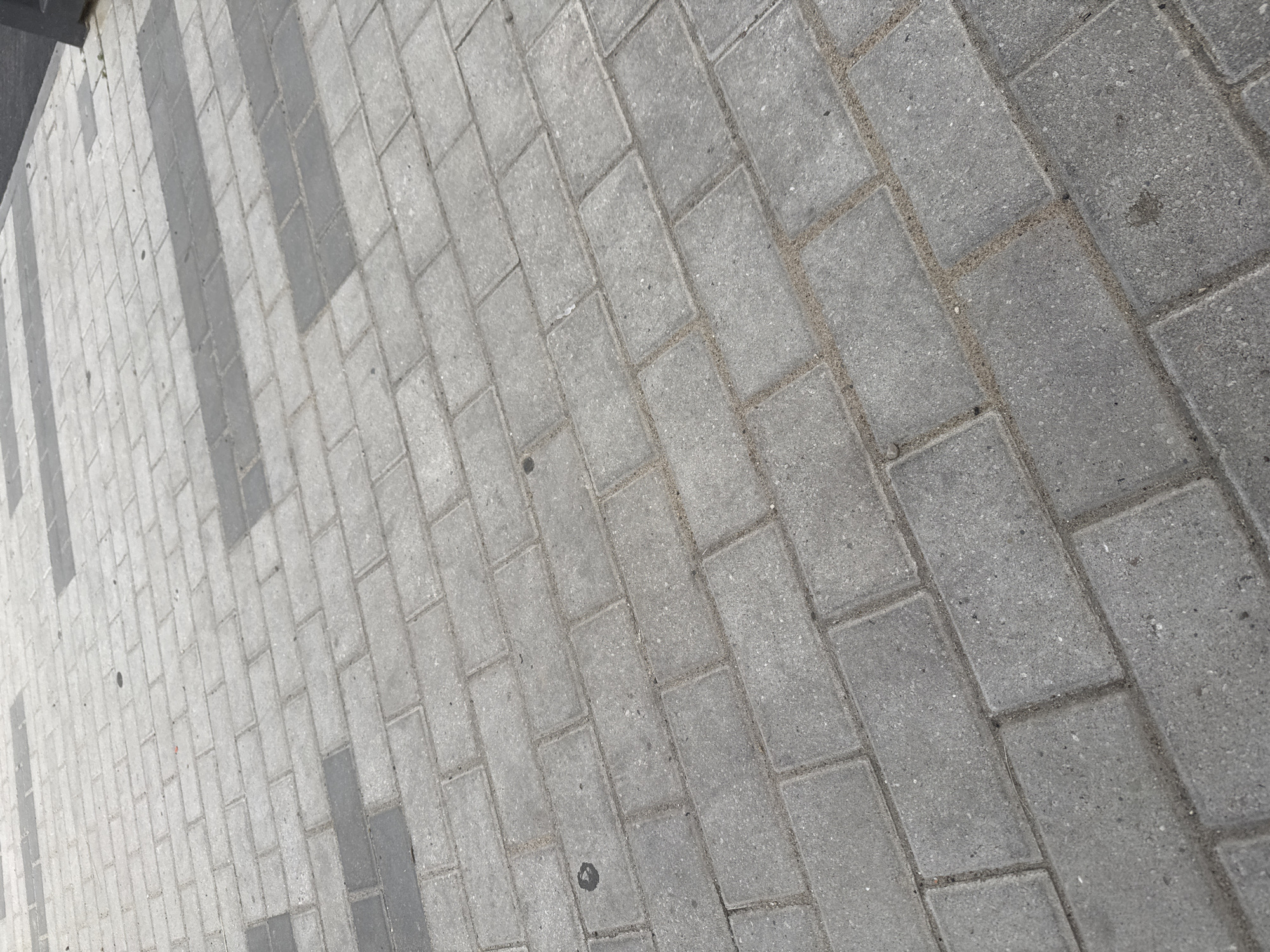 The image shows a section of a paved walkway in Koltsovo, Russia. The walkway is constructed from rectangular interlocking concrete pavers arranged in a running bond pattern. The pavers vary in color, with some being a lighter gray and others a darker gray, forming a subtle striped design across the walkway. The joints between the pavers are filled with sand. There is a dark rectangular object, possibly a bollard or a portion of a structure, visible along the right edge of the frame. The walkway appears to be outdoors, with no identifiable buildings or significant vegetation in the visible area. The lighting suggests it is daytime.