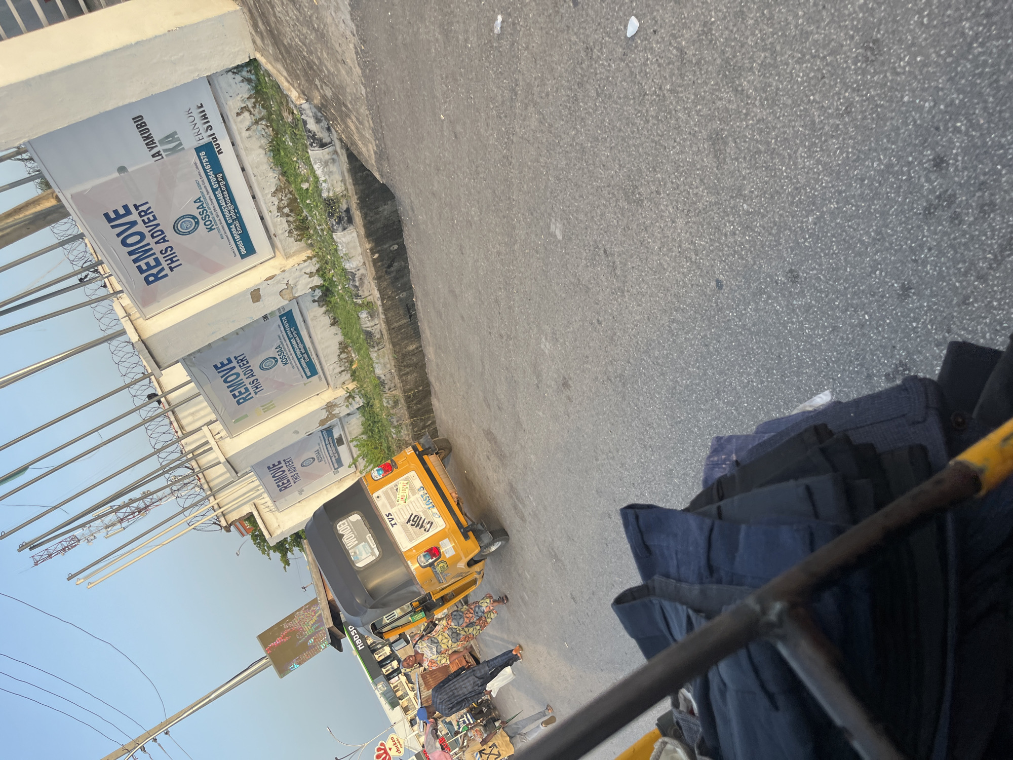 The image shows a street scene in an urban environment. In the foreground, a person's perspective from within a vehicle or cart reveals a stack of dark blue fabric. The street is paved with asphalt, and there are patches of dry grass and a concrete gutter along the side. In the middle ground, a yellow auto-rickshaw, also known as a Keke Napep, is parked. The auto-rickshaw has a black roof and displays a license plate with the characters "AU 10290". A woman in a brightly colored dress is standing near the auto-rickshaw, and a man in a blue patterned shirt and dark pants is walking away from it, carrying a white plastic bag. Further in the background, there are buildings with white facades that have large posters or advertisements on them. These advertisements appear to be political or public service announcements, with prominent text like "REMOVE THIS ADVERT" and mentions of "KOSSA" and a governor's name. Multiple flagpoles are also visible, some with flags. The sky is clear and appears to be late afternoon or early evening, casting long shadows. The overall atmosphere suggests a busy street in a Nigerian city.