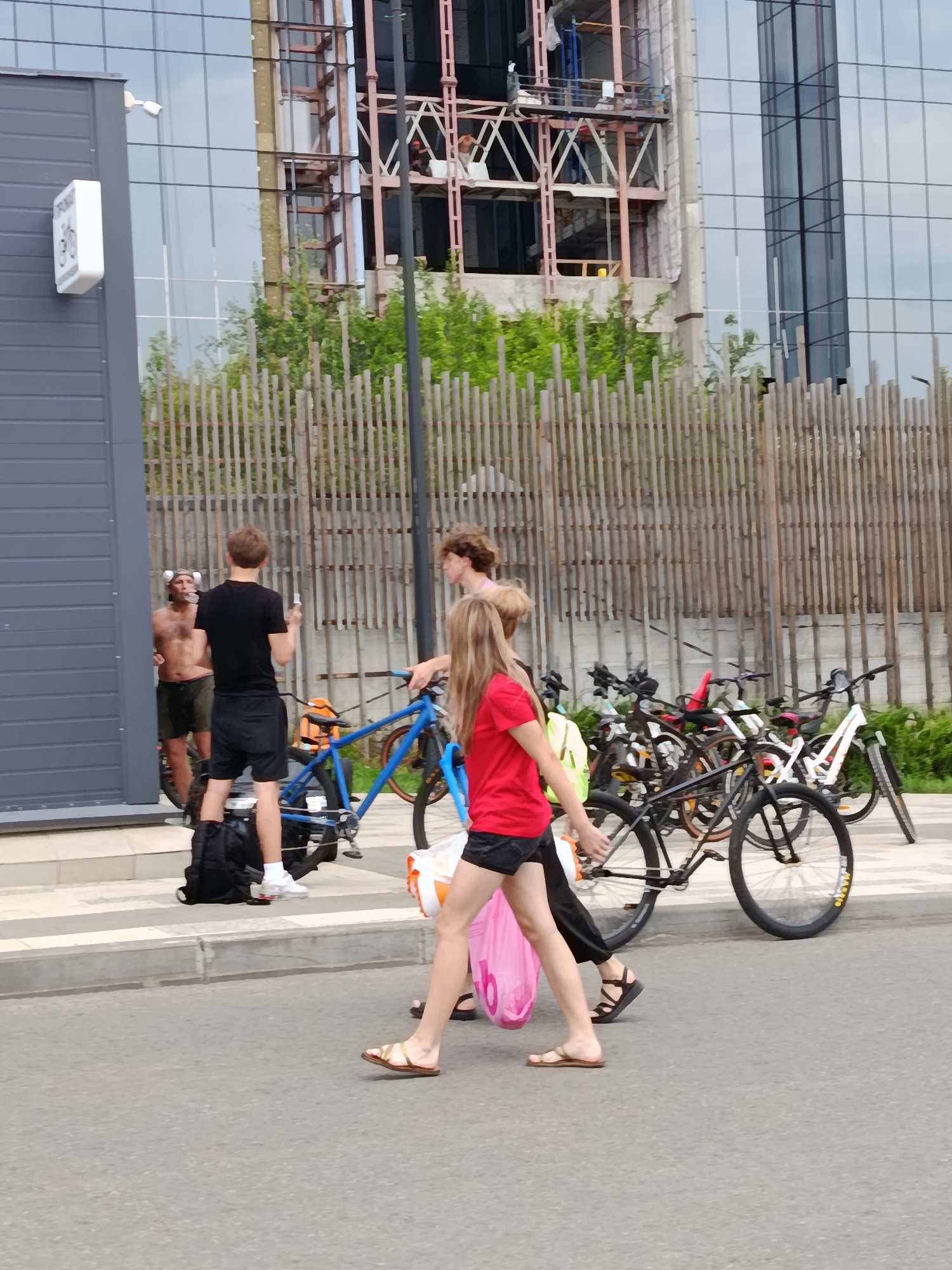 The image depicts a street scene in Saratov, Russia, in front of a modern building with a glass facade, part of which appears to be under construction. A wooden fence runs horizontally across the mid-ground.
In the foreground, on the pavement, two young women are walking from left to right. The woman on the left is wearing a red t-shirt and black shorts, carrying a pink plastic bag. The woman on the right is wearing black trousers. A group of bicycles is parked behind them.
To the left of the women, a young man in a black t-shirt and shorts stands holding a white object. To his left, a man with no shirt on and dark shorts is leaning against a dark blue bicycle, holding a cigarette to his mouth. Another man with curly hair is visible behind the man in the black t-shirt. A black backpack is on the ground near the man in the black t-shirt.