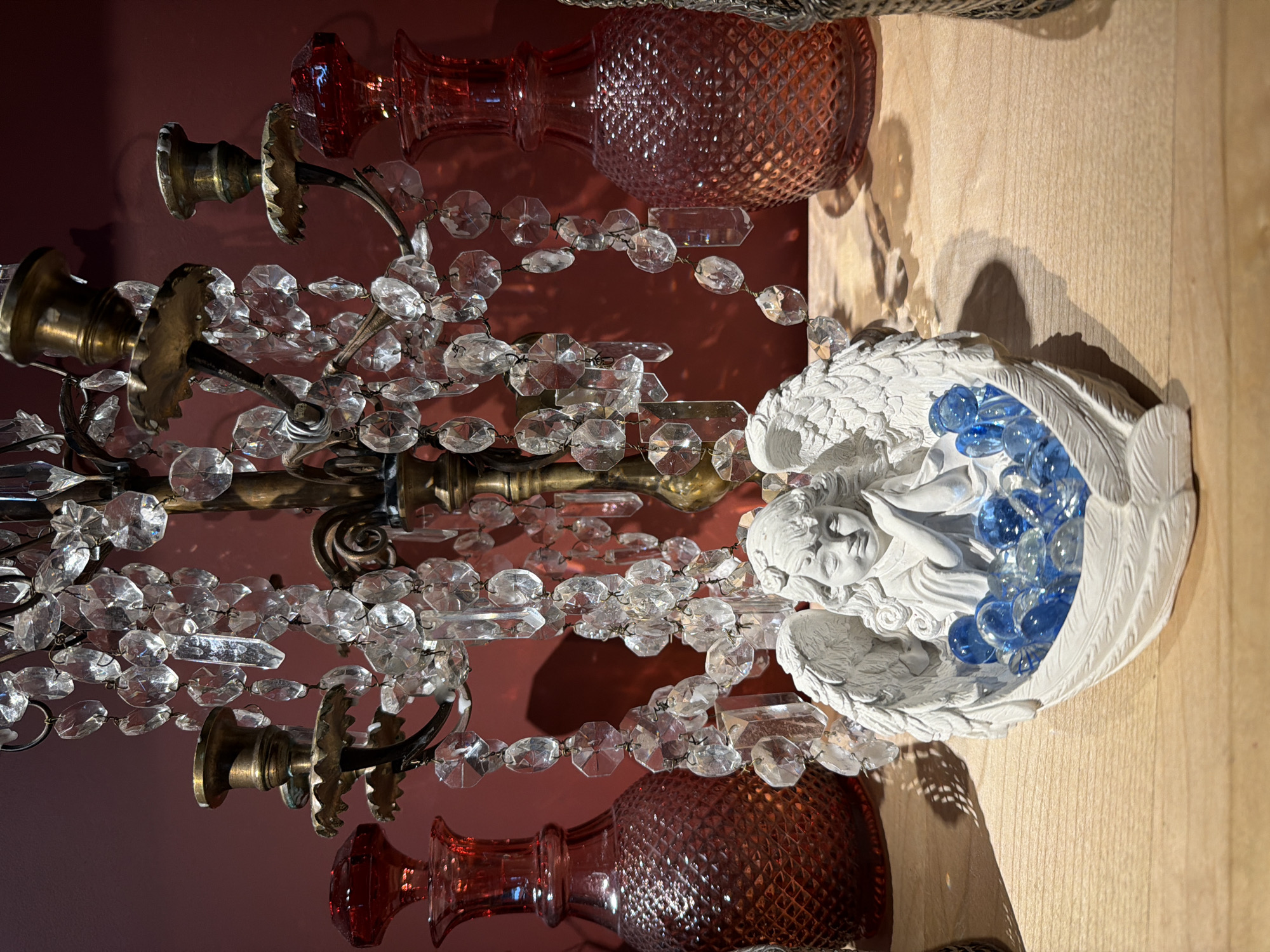 The image shows a still life arrangement. There is a brass candelabra with crystal bead strands hanging from it. The candelabra has three arms, each with a candle holder. Two red glass vases with a textured pattern are also in the scene. An angel figurine, white in color and holding blue marbles, is positioned to the right of the candelabra. The background is a red wall. The scene is situated on a light-colored wooden surface.