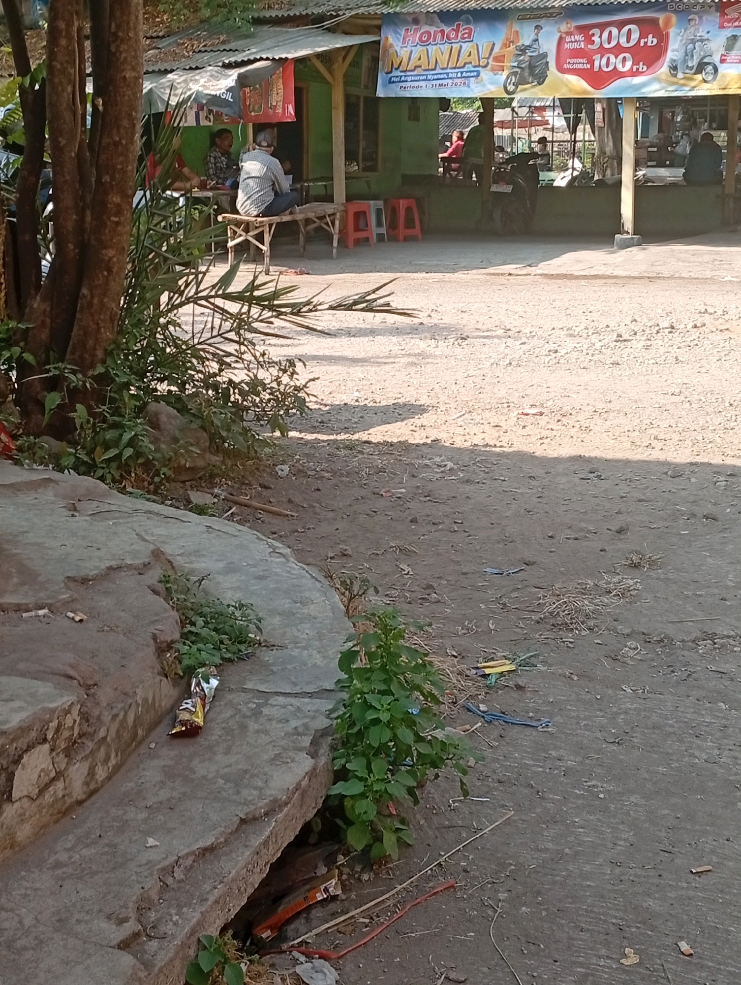 The image depicts an outdoor scene in Jogosari, Indonesia, near a small roadside establishment. A banner advertising "Honda MANIA!" with prices and financing options is prominently displayed. Several individuals are seated at tables outside the establishment, appearing to be customers or patrons. A motorcycle is parked near the building. The ground is a mix of dirt and gravel, with some scattered debris including cigarette butts and wrappers. A concrete structure with some overgrowth is visible in the foreground. The overall impression is of a casual, possibly rural or semi-urban setting.