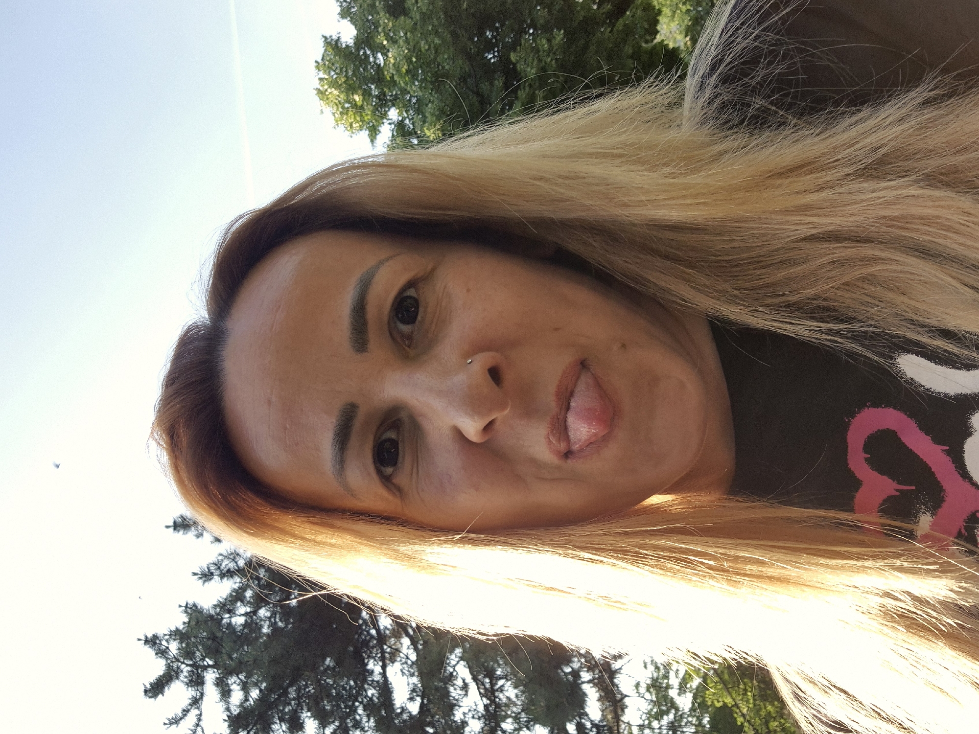 A woman with blonde hair is making a face at the camera, sticking out her tongue. She has a nose piercing and is wearing a black t-shirt with a pink and white design. The background consists of trees and sky, suggesting an outdoor location.